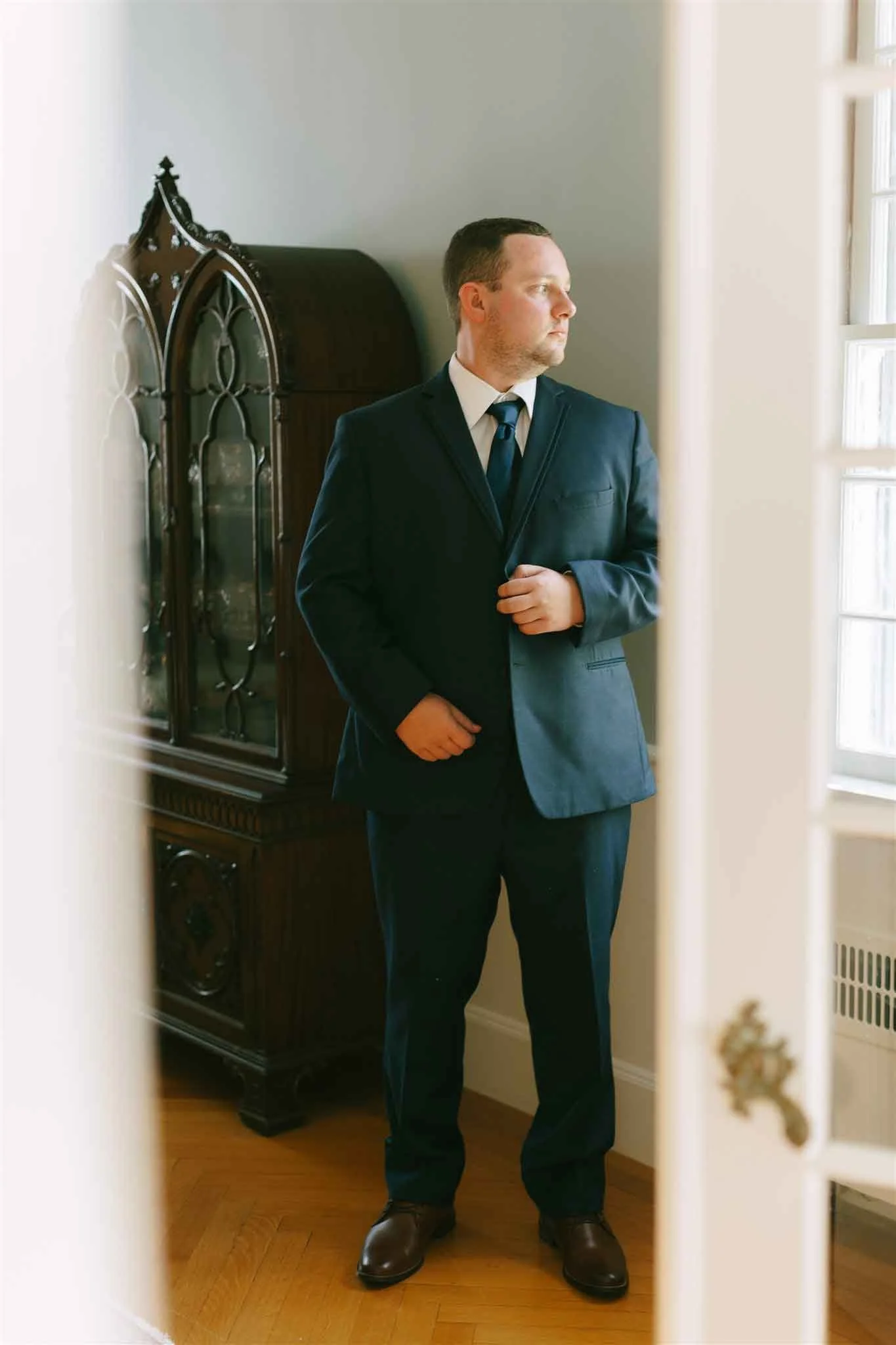 A man in a suit standing indoors, looking out a window, photographed through a partially open door.