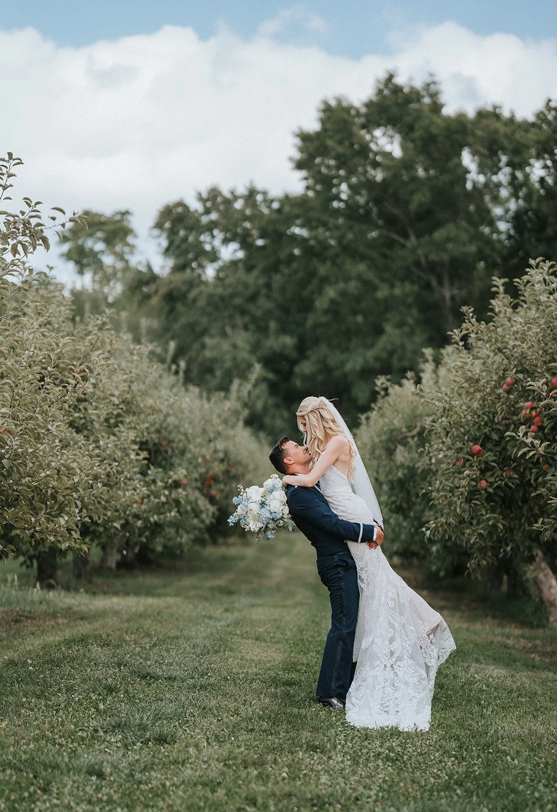 A newlywed couple in wedding attire, with the groom lifting the bride in an orchard, surrounded by green trees and grass.