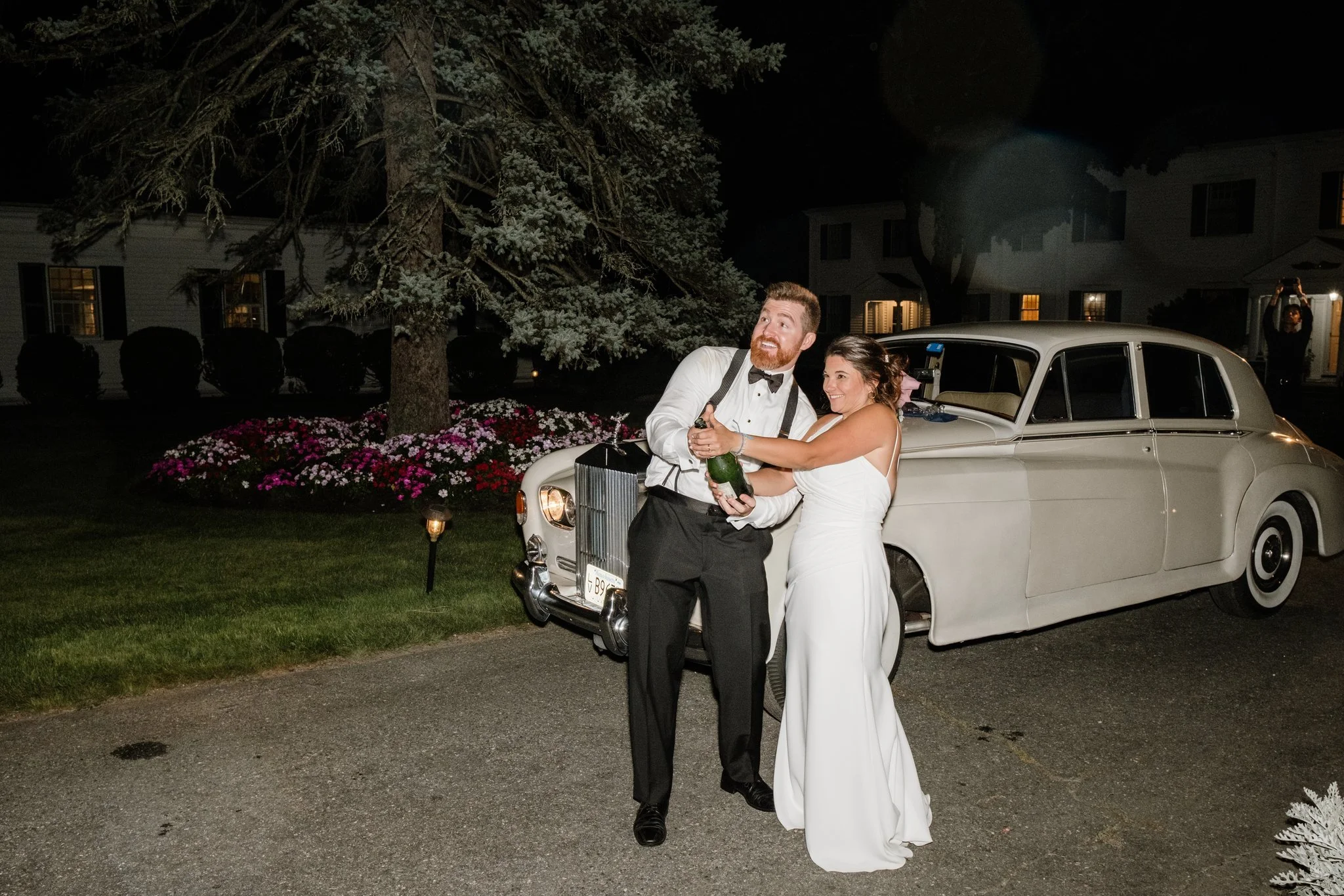 A couple in formal wedding attire celebrating outside at night. The man is holding a champagne bottle, and the woman is smiling and holding onto it. Behind them is a vintage white car and a landscaped garden with flowers and a large tree.