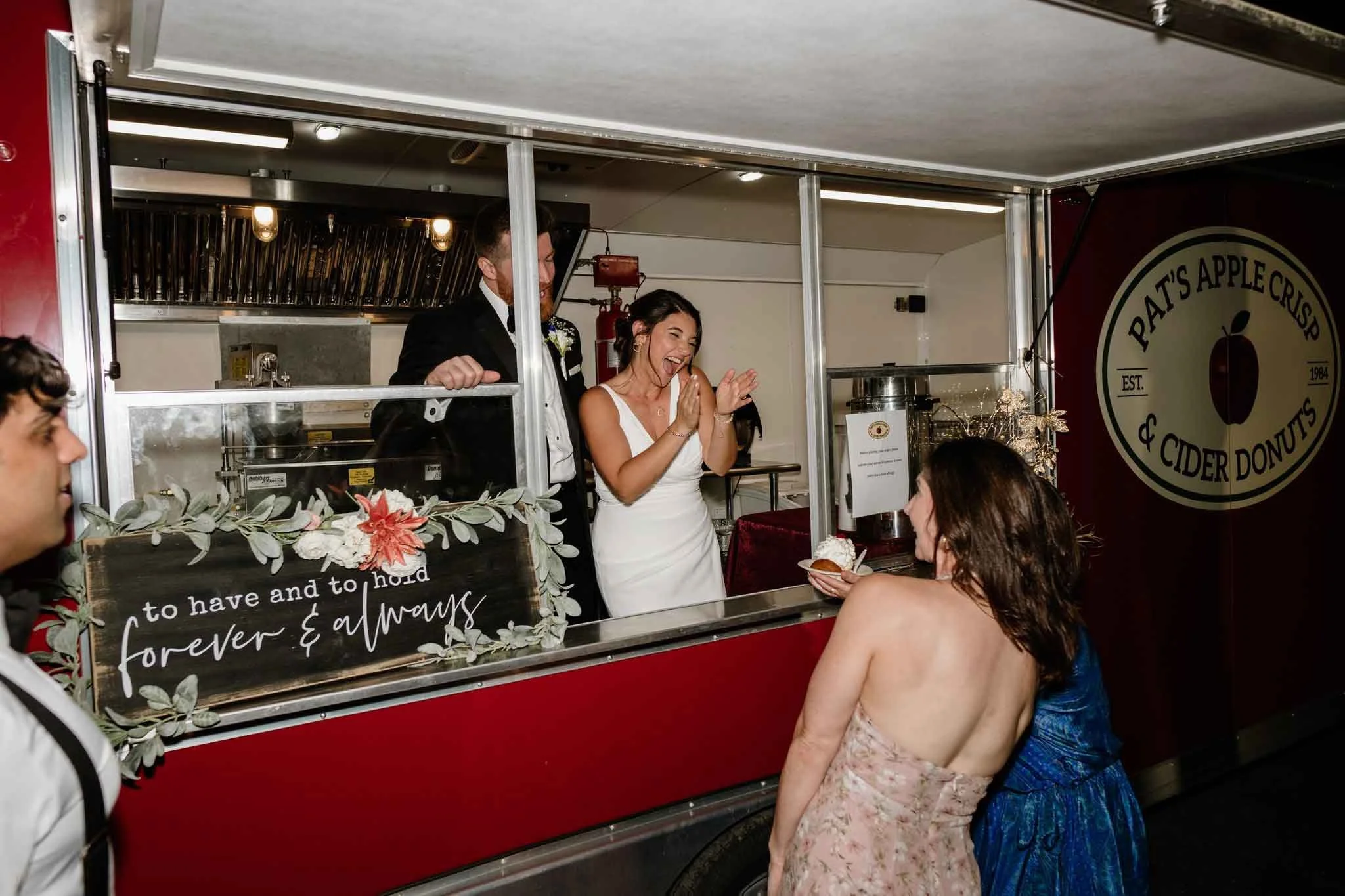 A bride in a white wedding dress is smiling and clapping while standing at a food truck serving ice cream or dessert to women in dresses, one in a floral dress and the other in a blue dress. The food truck has a sign that reads "Pat's Apple Crips & C