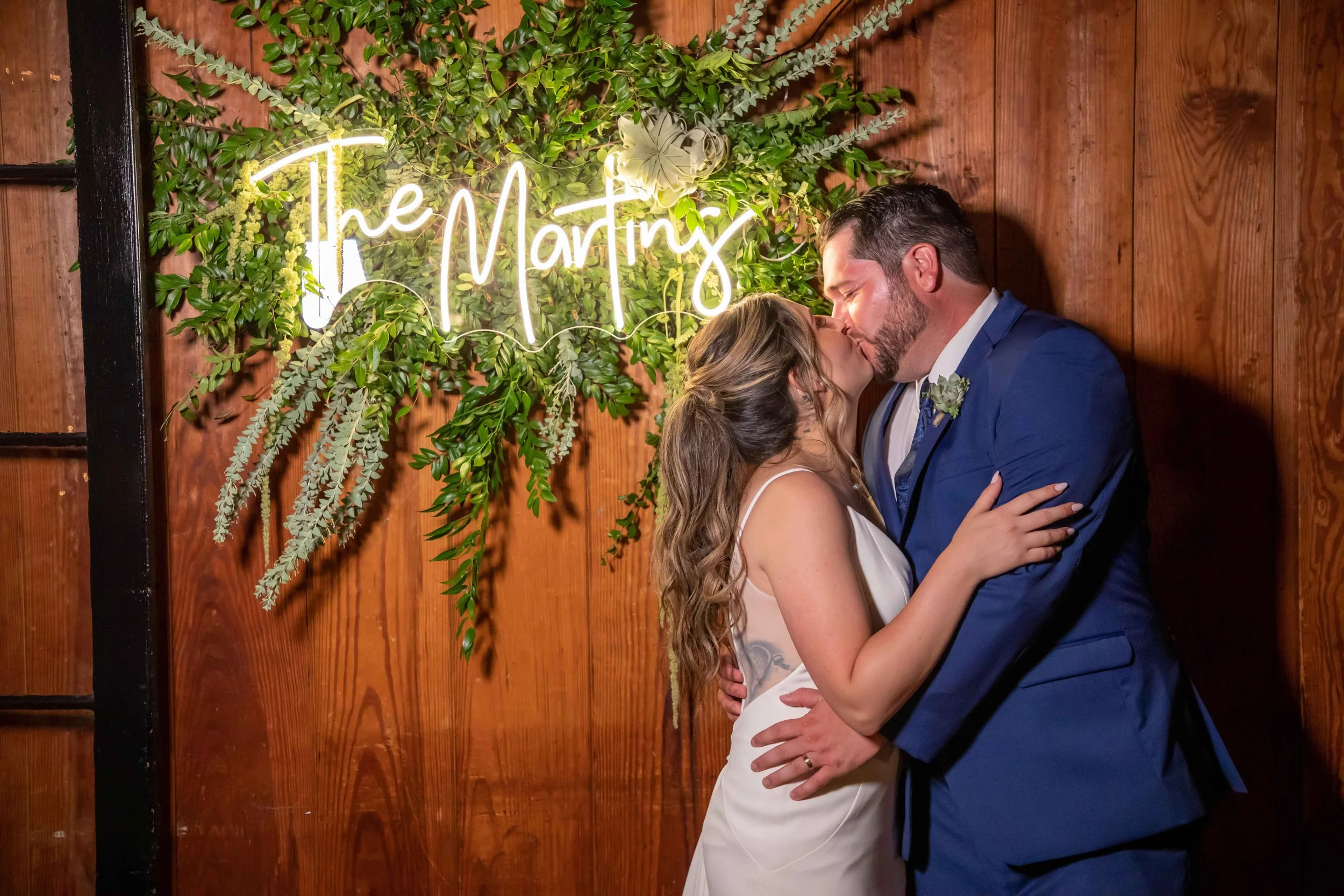 A couple kissing at a wedding, standing in front of a wooden wall with a green leafy decoration and neon sign that says 'The Martins'.