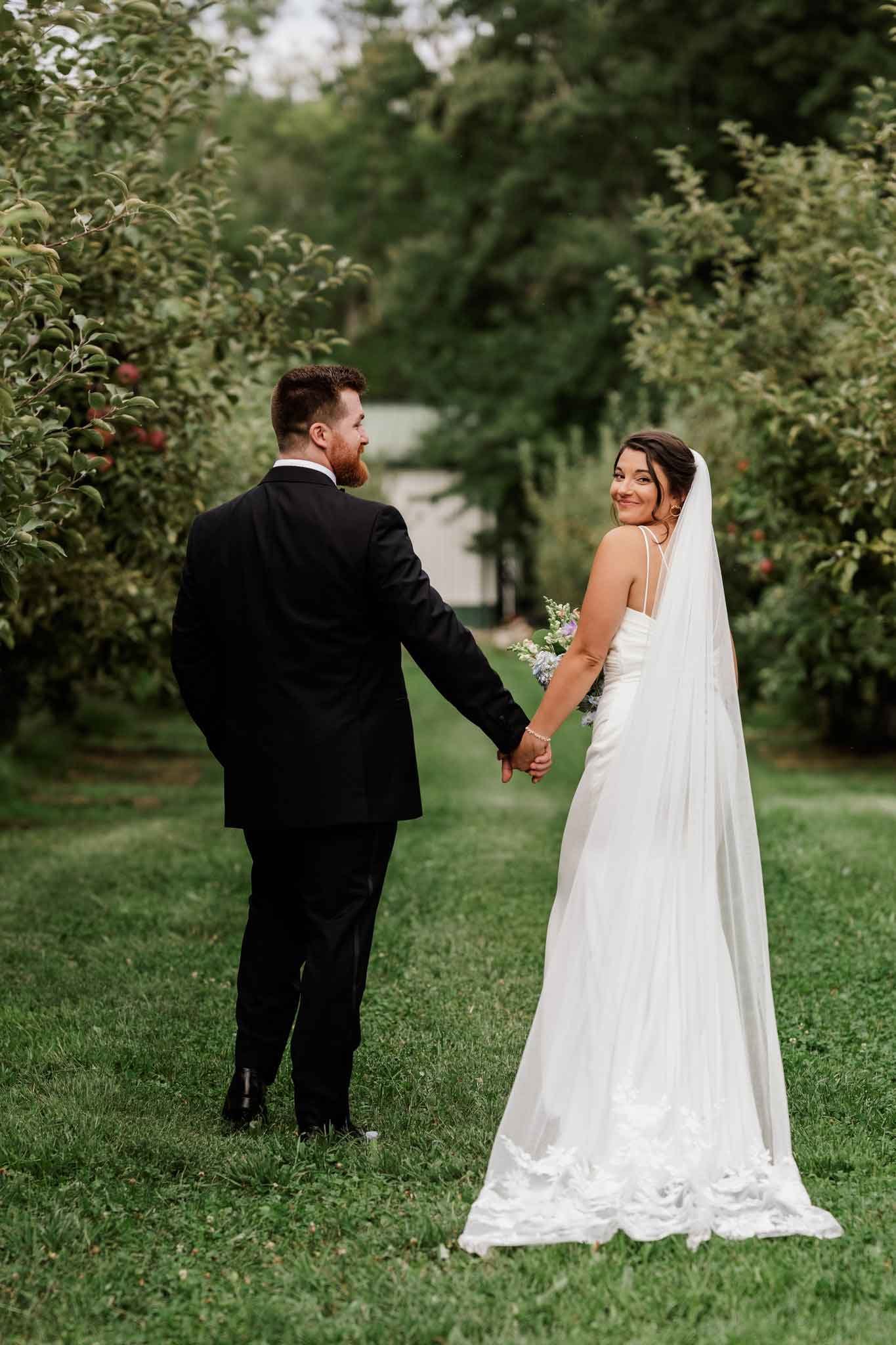 A bride and groom holding hands in a garden, with the bride in a white wedding dress and veil, and the groom in a black tuxedo, smiling at each other.