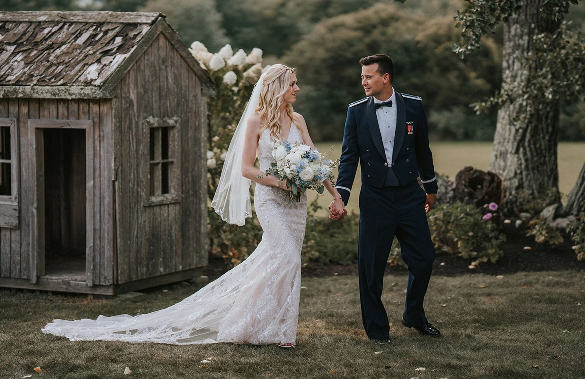 A bride and groom walking hand in hand outdoors, with the bride in a white wedding dress and veil holding a bouquet of white and blue flowers, and the groom in a military uniform, on a grassy area near a small wooden shed and trees.