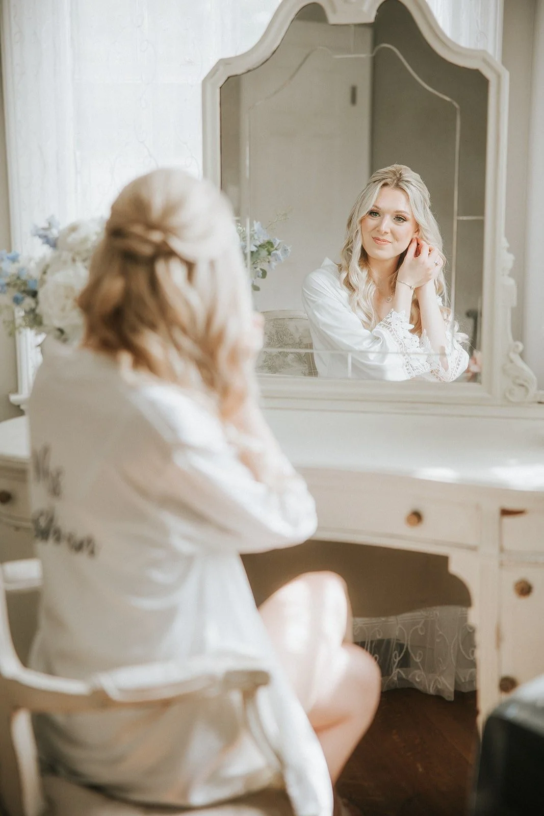 A woman with blonde hair sitting at a vintage white vanity table, adjusting her earring while looking at her reflection in the mirror, with flowers on the table.