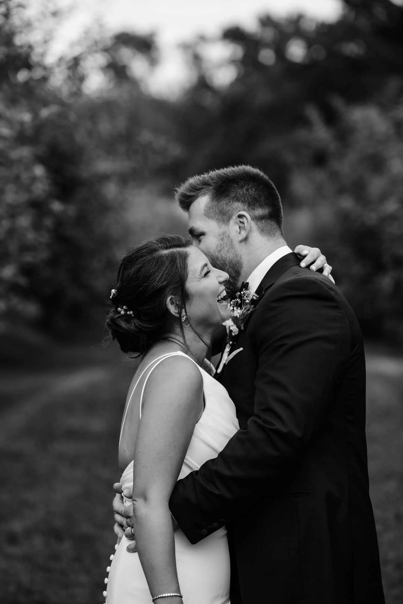 A black and white photo of a wedding couple sharing a tender moment outdoors, with the groom kissing the bride on her forehead. The bride is smiling and the background is blurred nature scenery.