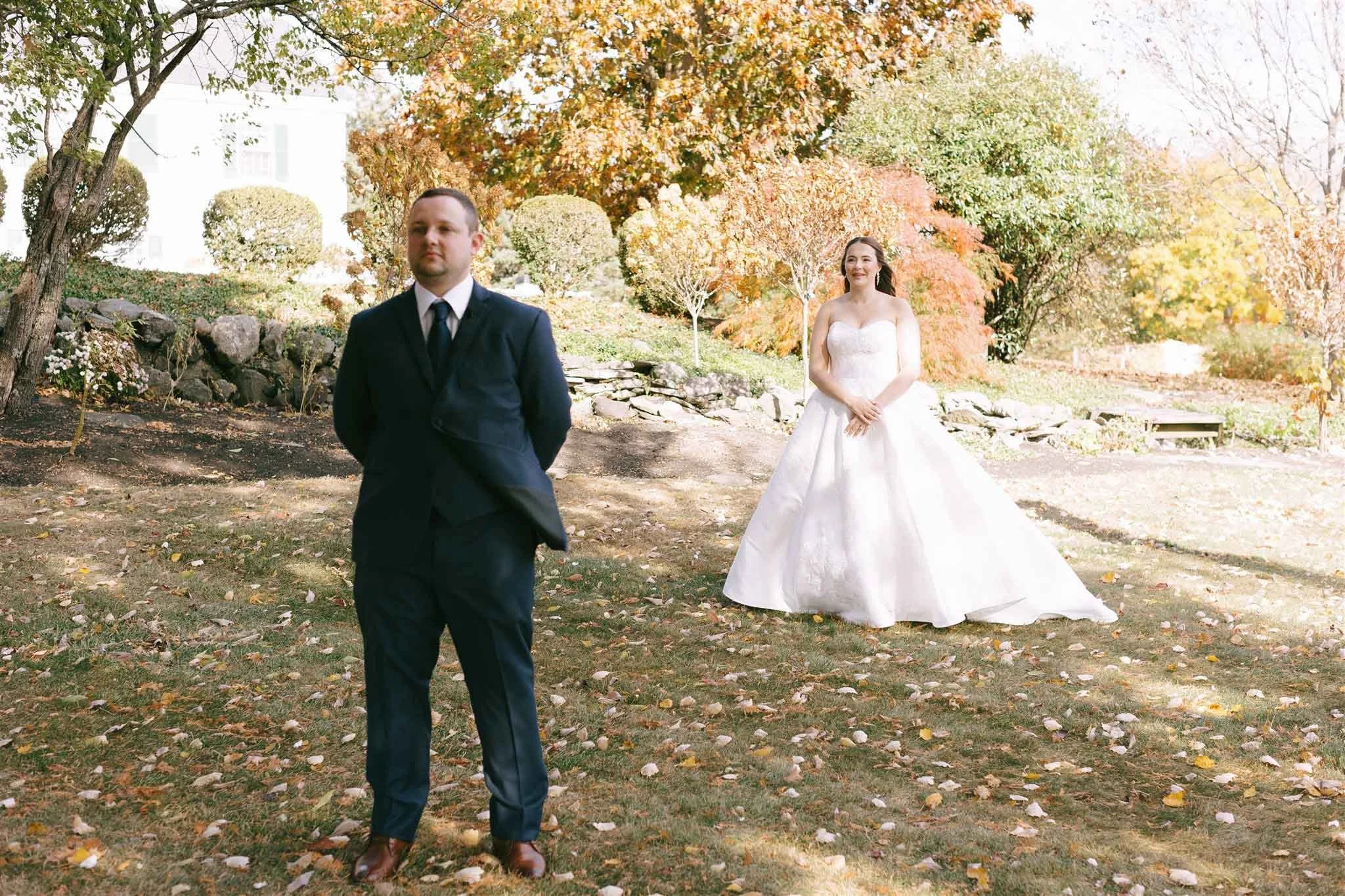 A man in a black suit stands with his hands behind his back in a park with fall foliage. A woman in a white wedding dress stands further back, smiling, among trees with orange and yellow leaves.