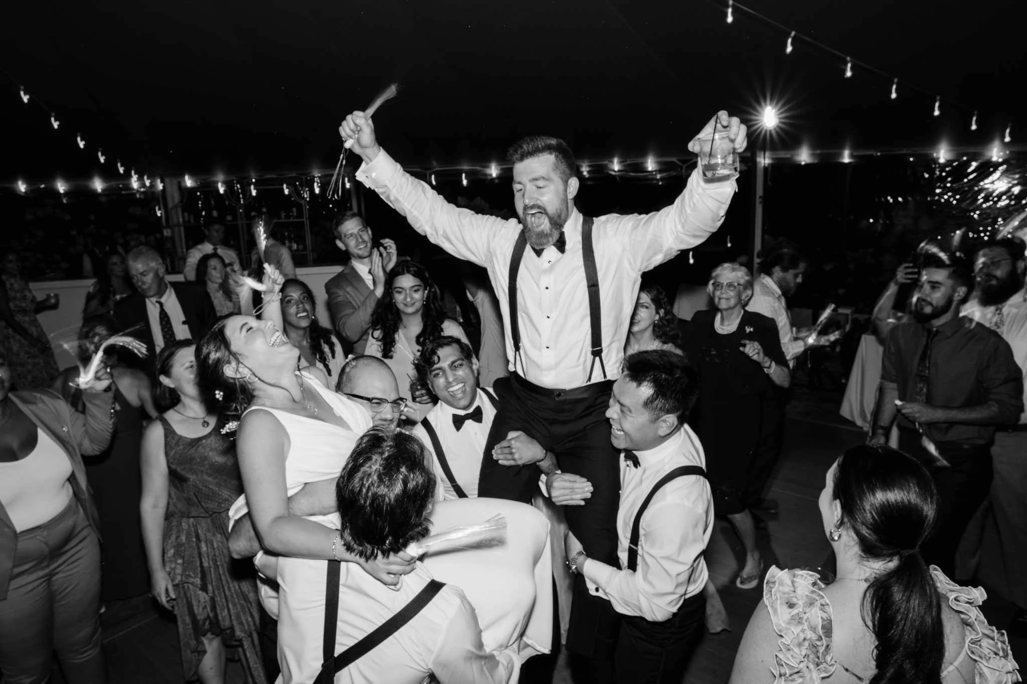 A lively celebration with a man being lifted by others, holding a sparkler and a drink, surrounded by smiling people at a party or wedding reception.