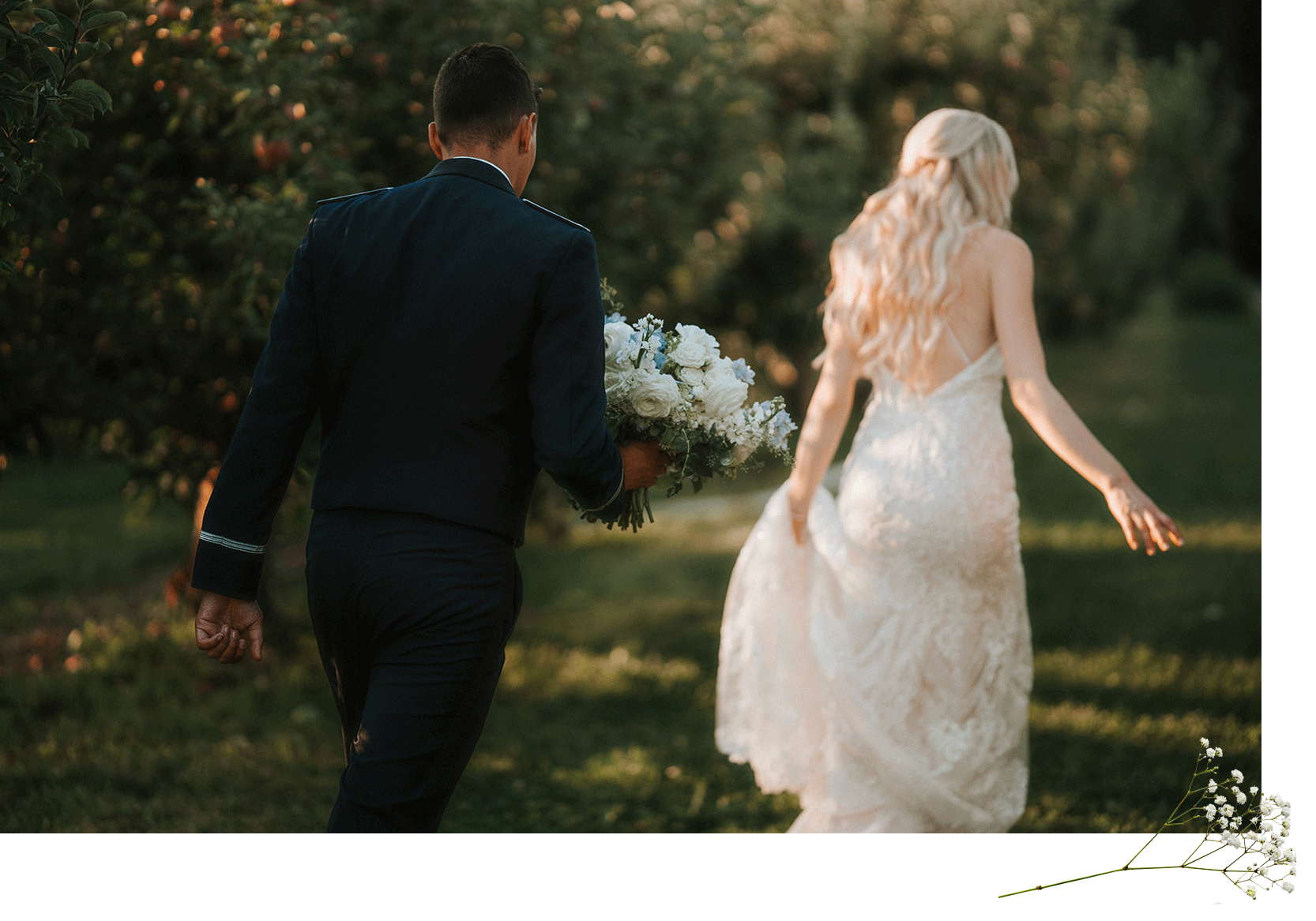 A man in a navy blue suit holding a bouquet of white and blue flowers walking behind a woman in a white lace wedding dress outdoors in a garden setting with sunlight filtering through trees.