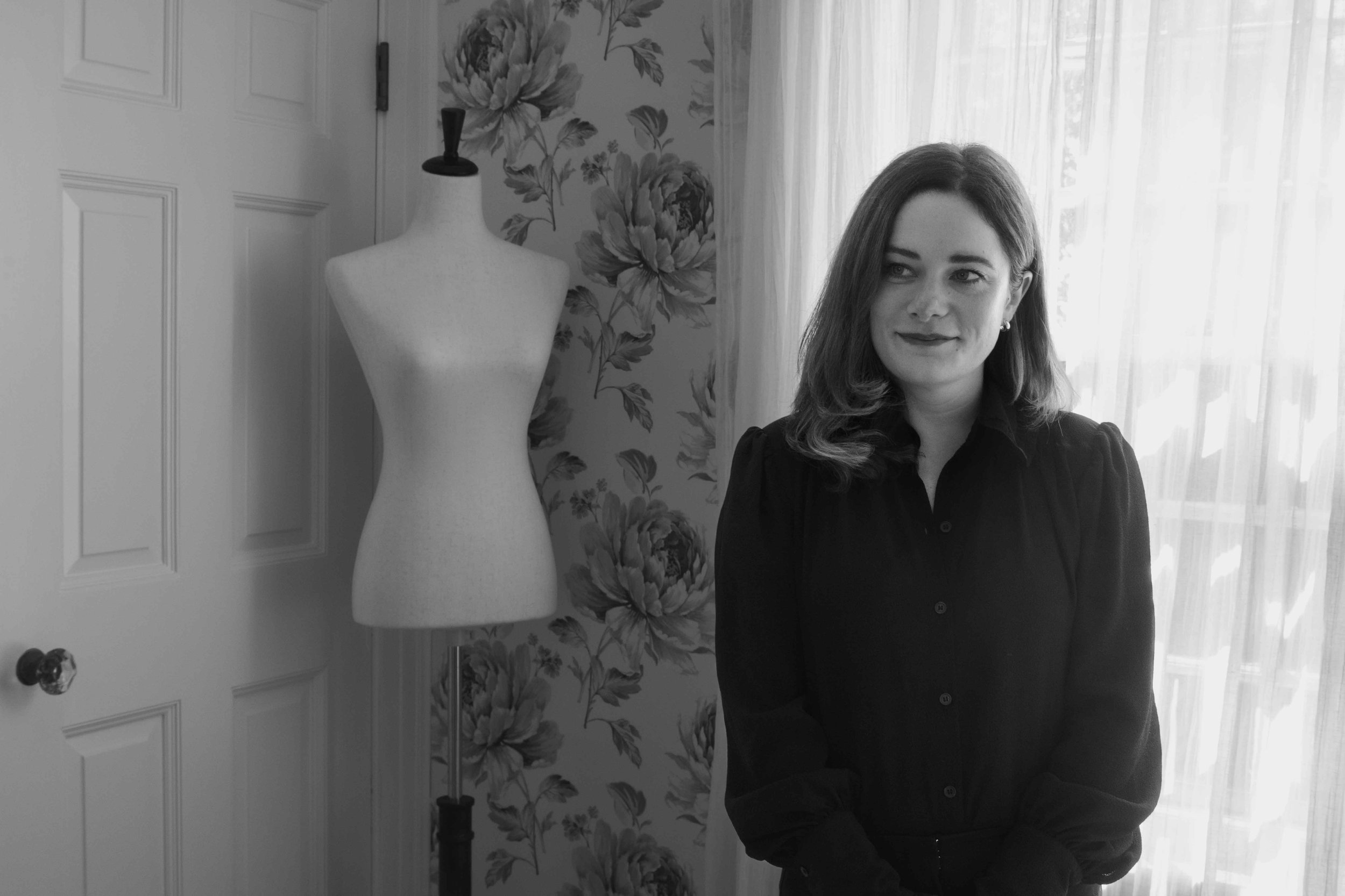 A woman with wavy hair standing in a room next to a dress form and floral wallpaper, with sheer curtains in the background.