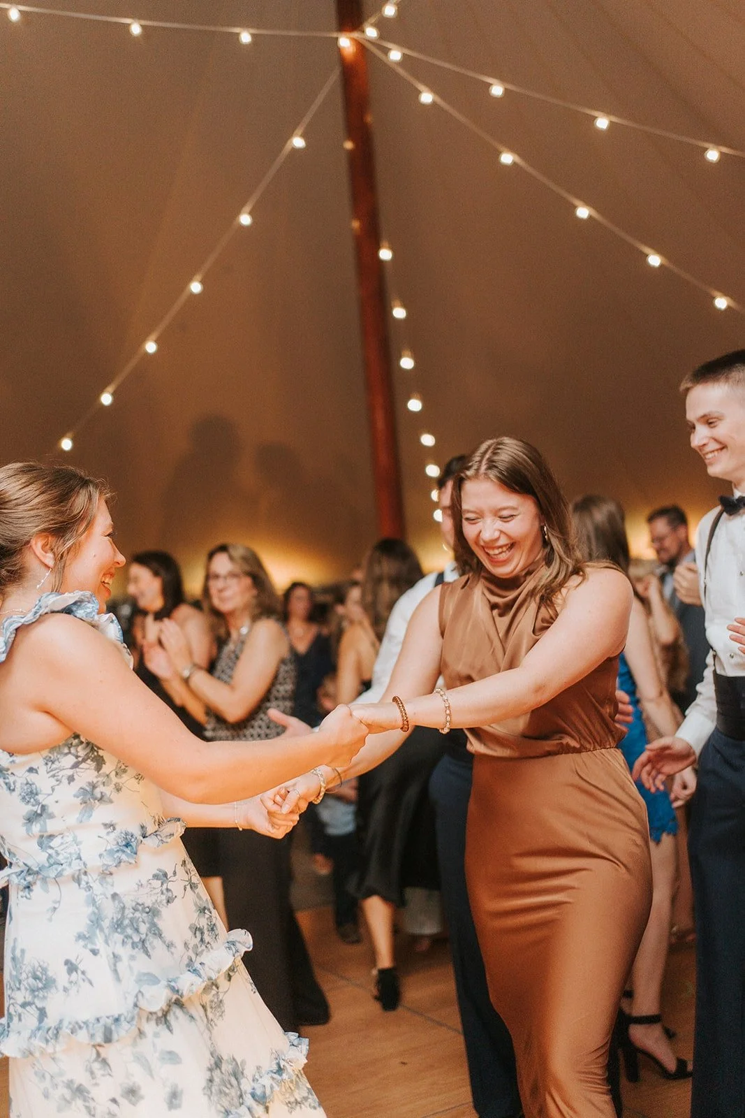 People dancing and laughing at a wedding reception under string lights in a tent.