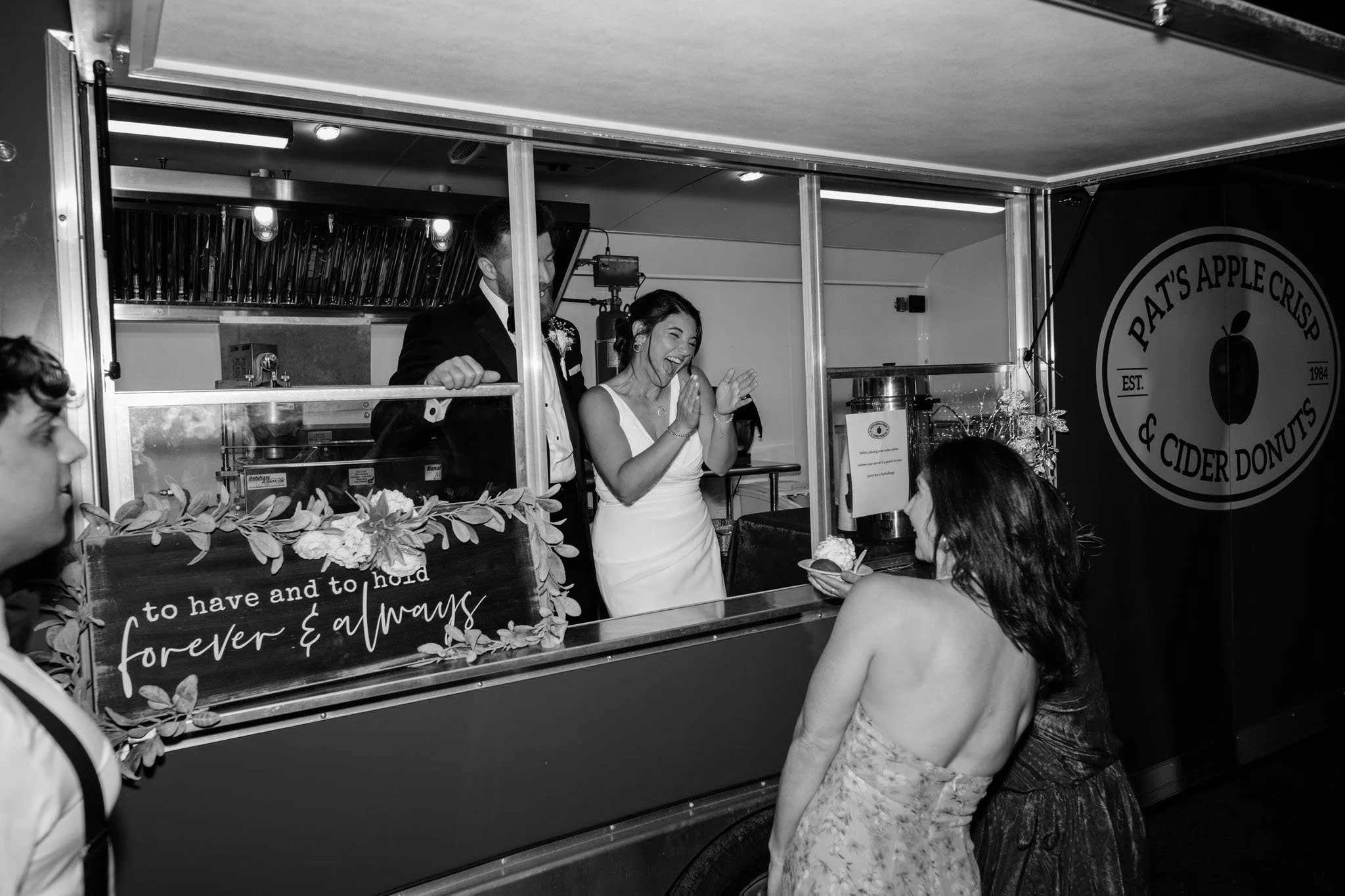 Bride and groom at a food truck wedding celebration, with a woman holding a dessert, smiling and clapping in front of the truck labeled 'Pat's Apple Crisp & Cider Donuts'.