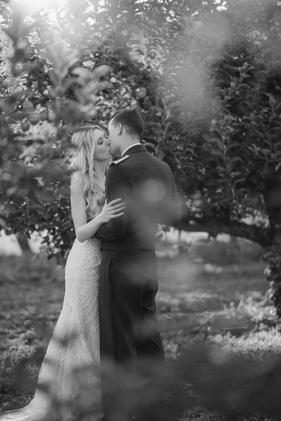 A couple dressed in wedding attire sharing a kiss in an orchard, with trees in the background and soft lighting.