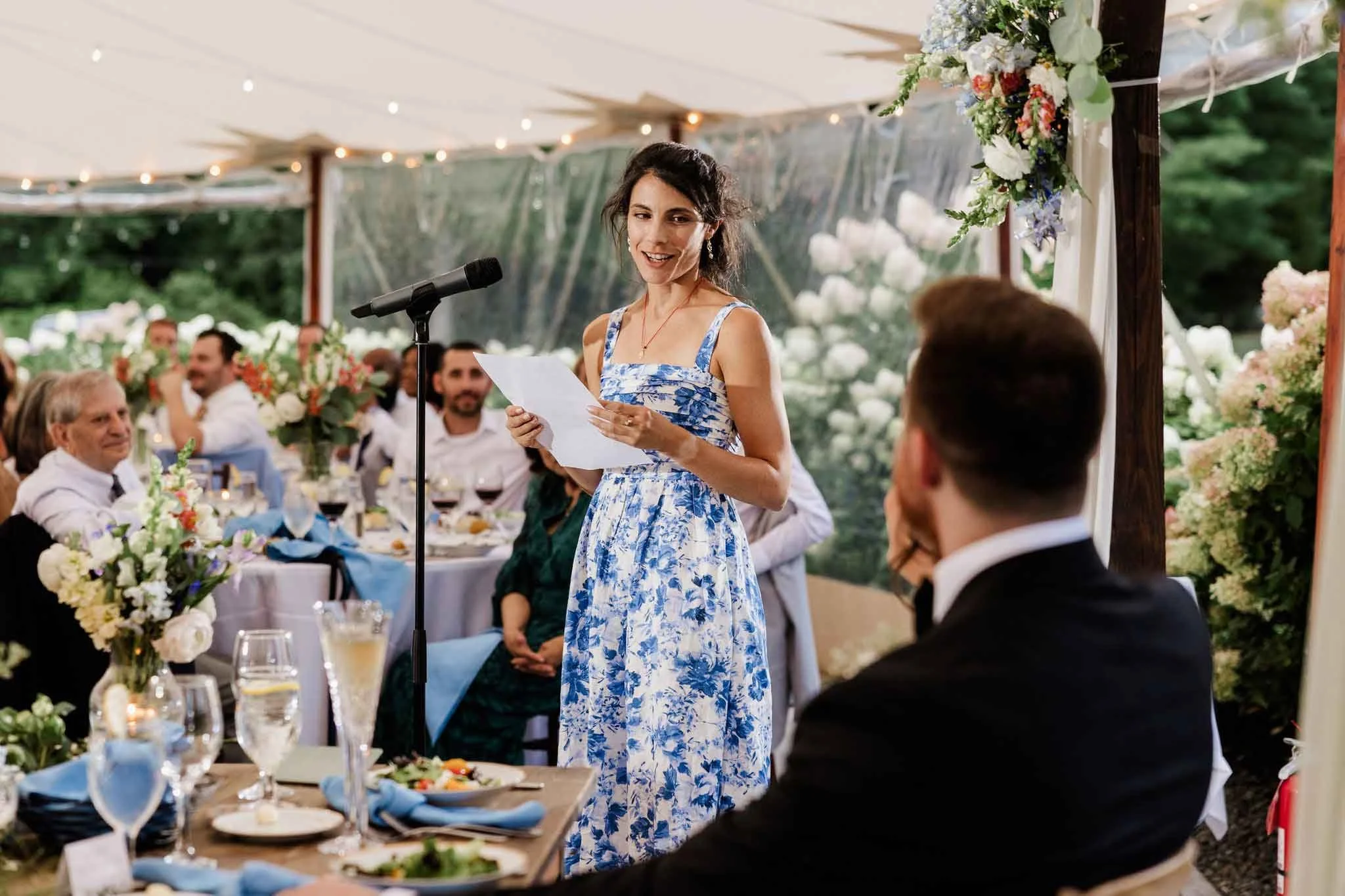 A woman in a blue and white floral dress giving a speech at a wedding reception under a decorated tent with guests seated at tables.