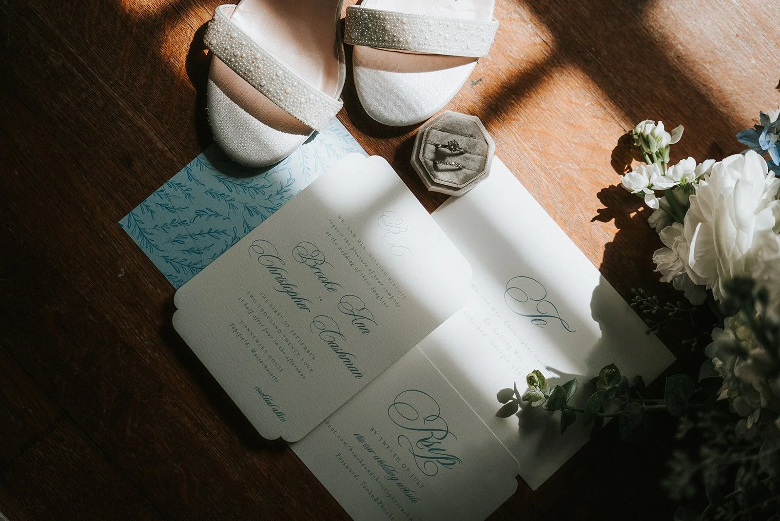 Wedding invitation, pearl-adorned white shoes, ring box with rings, white flowers, and greenery on a wooden surface with sunlight.