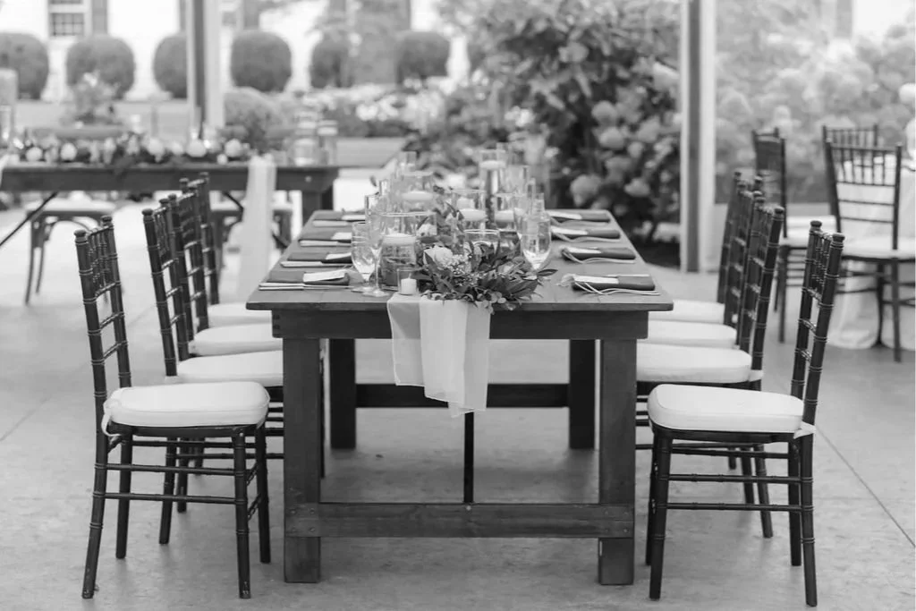 A long wooden dining table set with glasses, plates, and cutlery, decorated with a floral centerpiece and a table runner, in an outdoor setting.