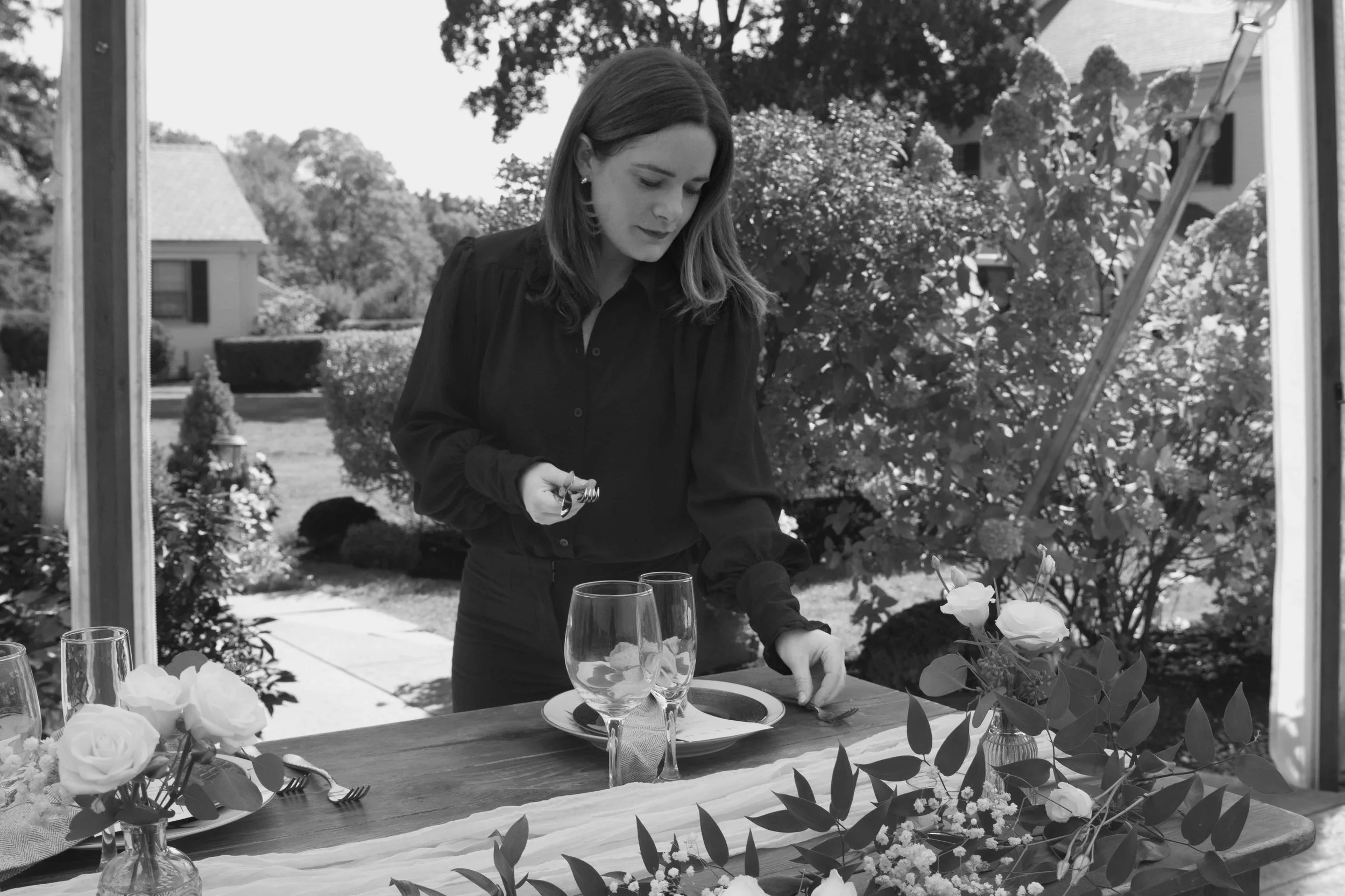 A woman setting up a table with flowers and glassware outdoors.