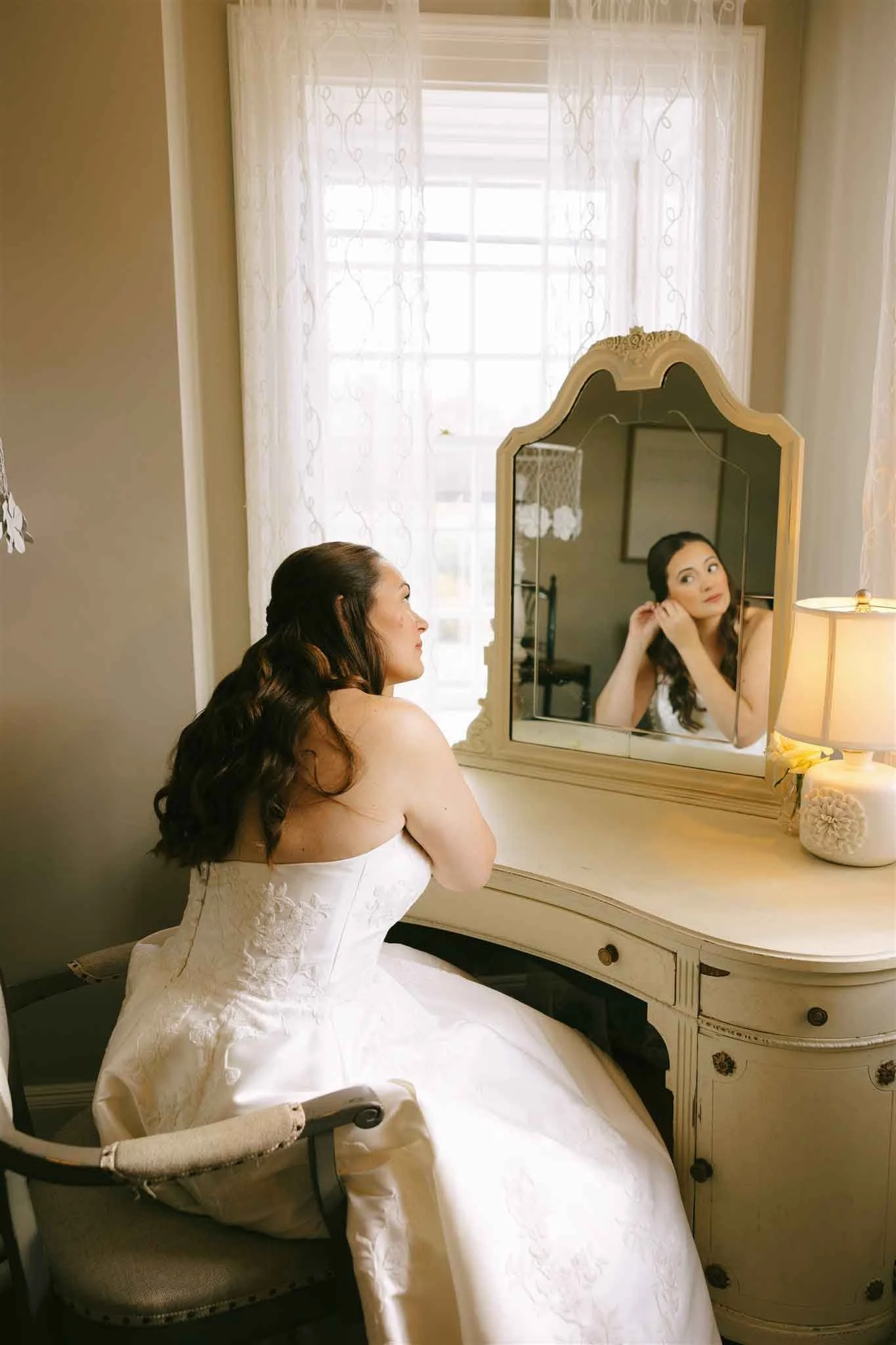 A bride sitting in front of a mirror, adjusting her earring, with a lit lamp on a dresser beside her.