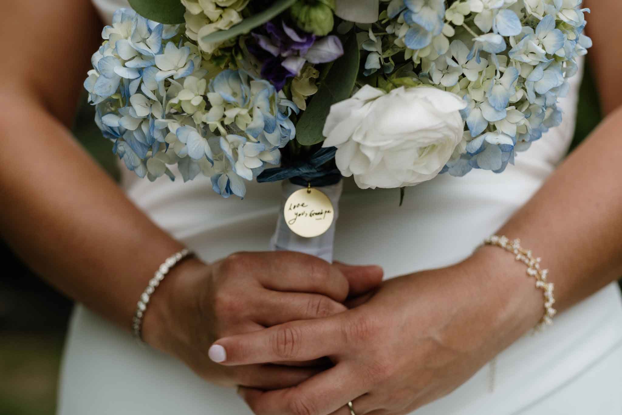 A person holding a bouquet of blue and white hydrangeas and white roses. The person is wearing bracelets on both wrists and a ring on the left hand. There is a small round tag on the bouquet with the message 'Love you, Grandma'.
