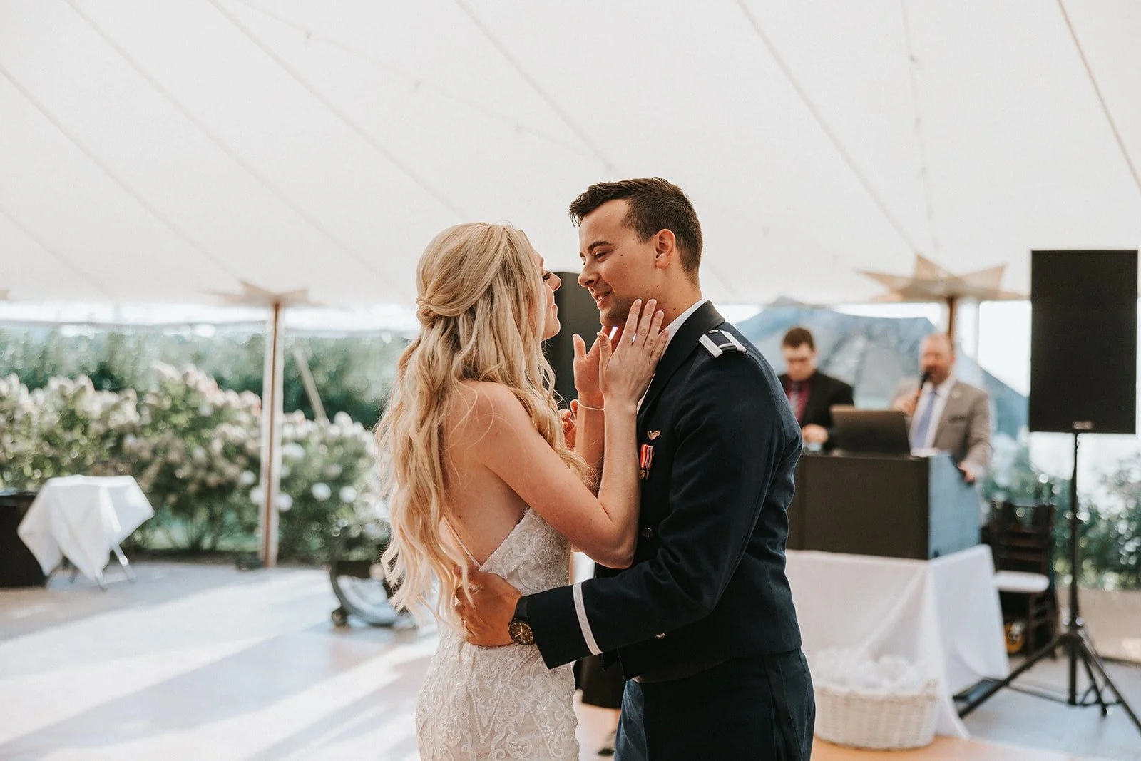 A bride and groom dancing at their wedding under a white tent, with a DJ and a speaker in the background.