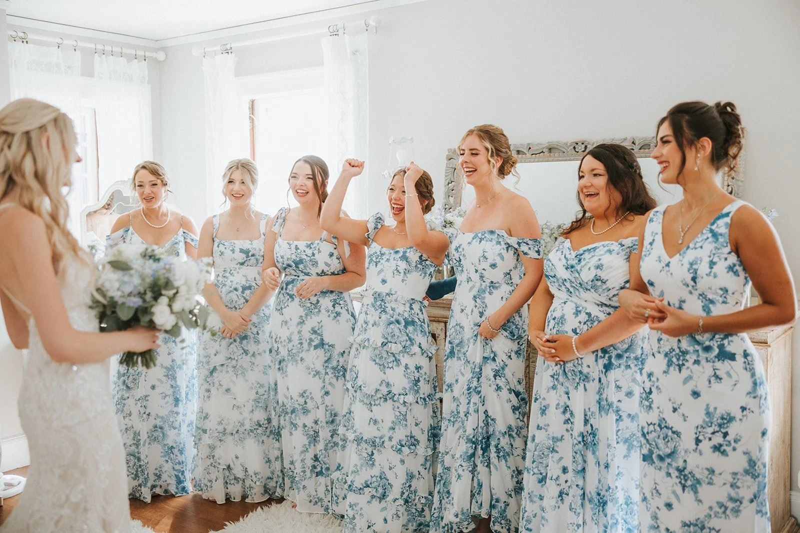 Group of women in matching blue and white floral dresses celebrating in a bright room with white curtains, with one woman holding a bouquet and smiling, while others are joyful, some with arms raised or clasped.