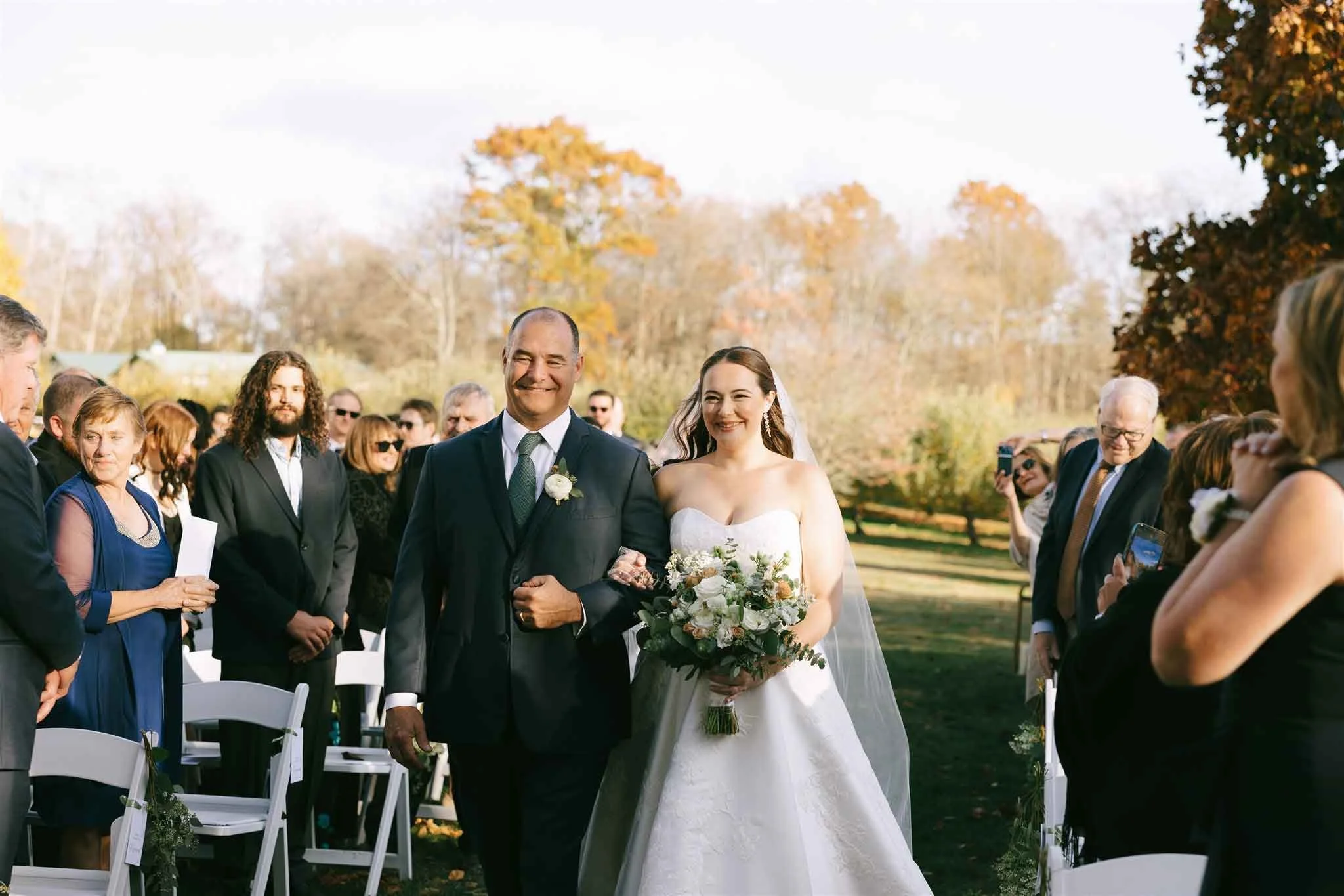 A bride walking down the aisle with her father during an outdoor wedding ceremony in autumn.