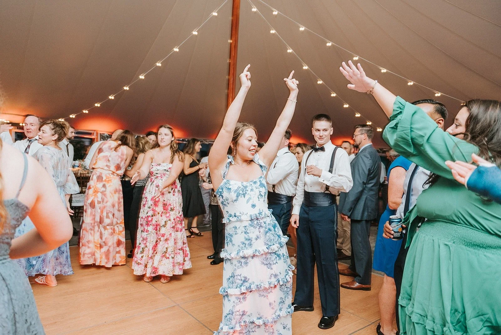 People dancing and celebrating at a party under a large tent decorated with string lights.