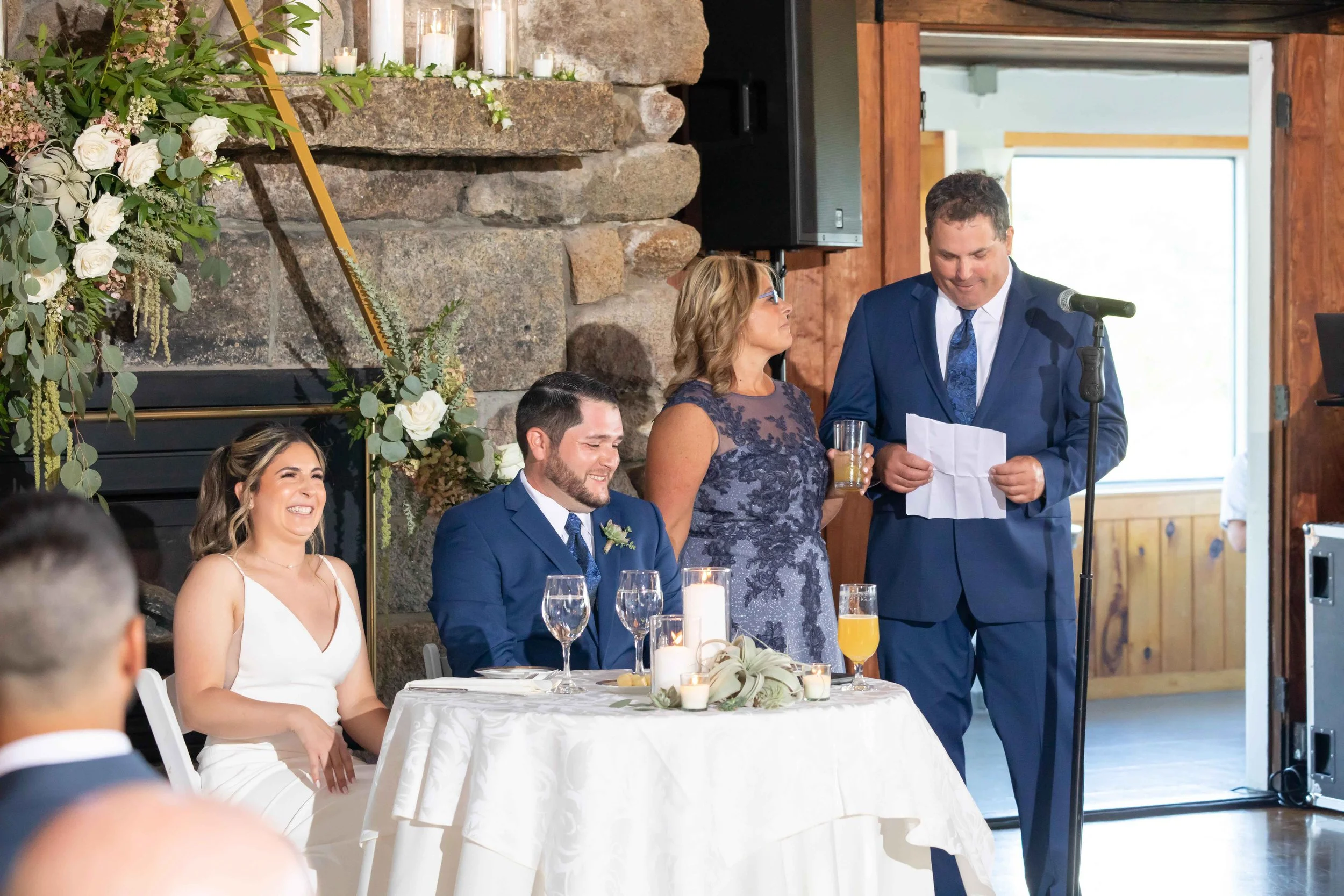 At a wedding reception, a bride and groom sit at a table, with a woman standing and speaking into a microphone. The couple is dressed in formal attire, with the bride in a white dress and the groom in a blue suit. The scene is decorated with floral a