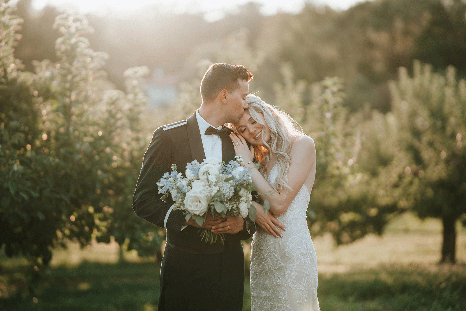 A bride and groom share a tender moment outdoors during their wedding, with the groom kissing the bride on the forehead as she smiles happily, holding a bouquet of white and green flowers.