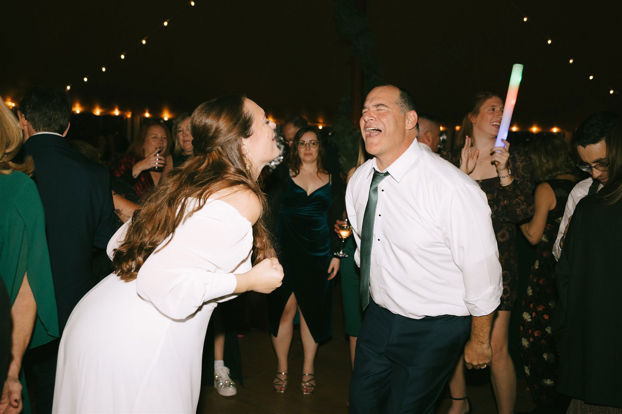 A woman and a man are dancing and singing together at a party, surrounded by other guests with string lights overhead.