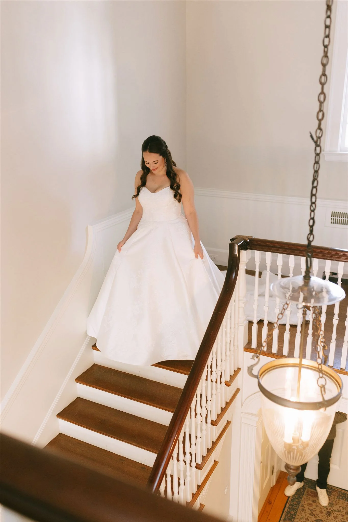 A woman in a white wedding dress standing on a staircase, looking down and holding her dress.