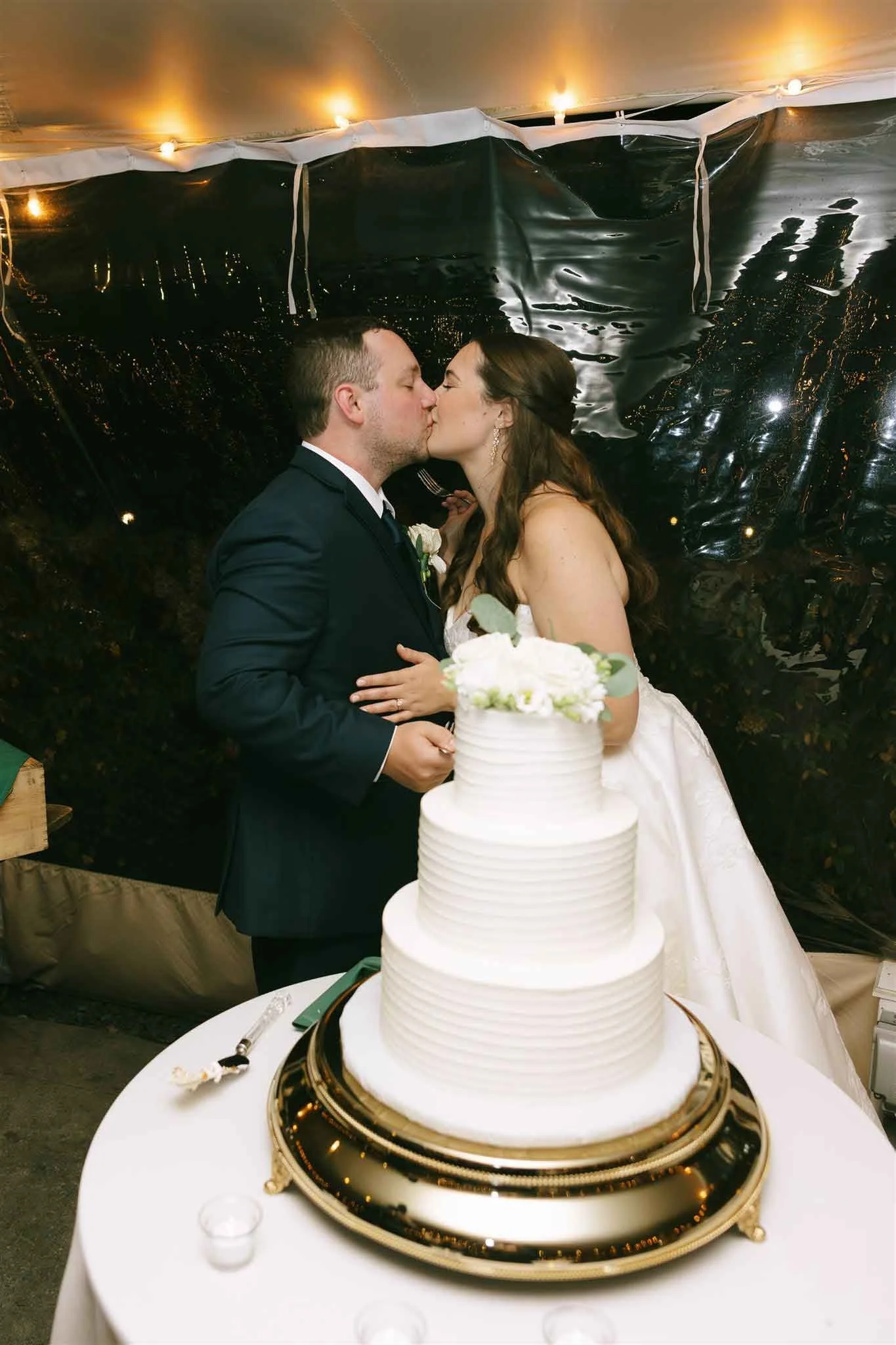 A bride and groom kiss at their wedding reception, standing behind a three-tiered wedding cake on a table with a white tablecloth.
