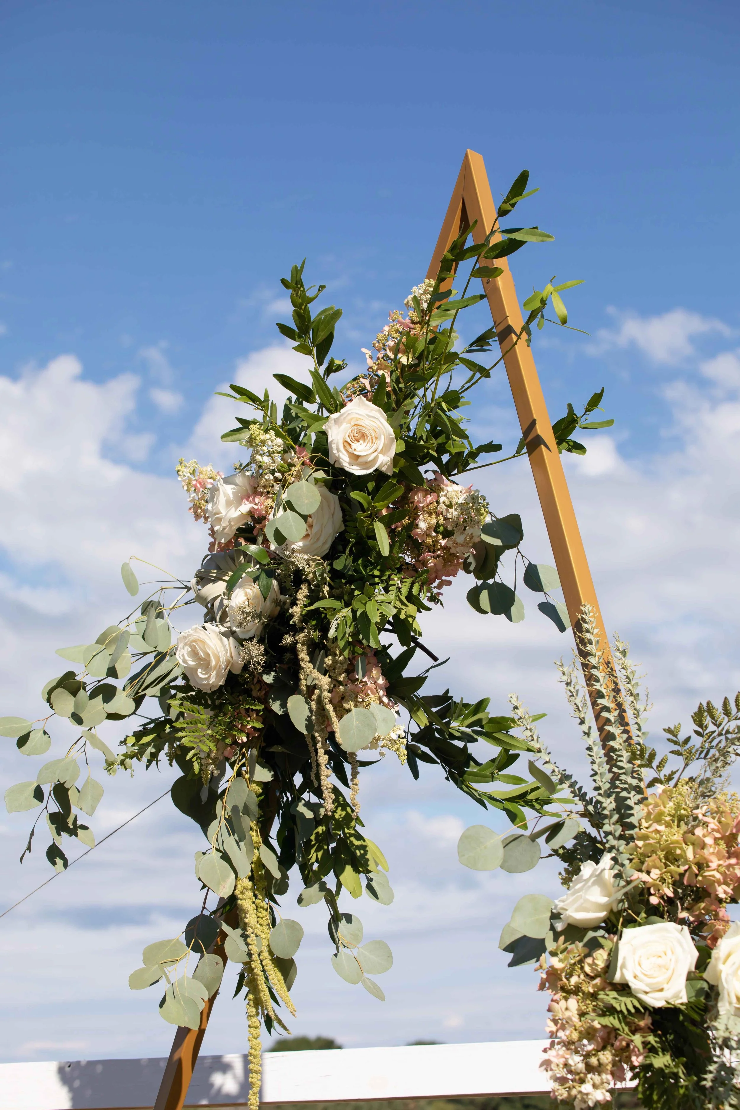 Outdoor wedding arch decorated with white roses, greenery, and small flowers against a blue sky with scattered clouds.
