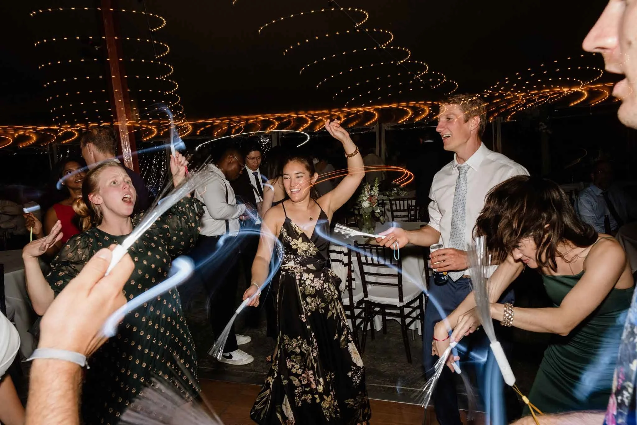 People dancing and celebrating at a party or wedding reception with sparklers and colorful lights overhead.