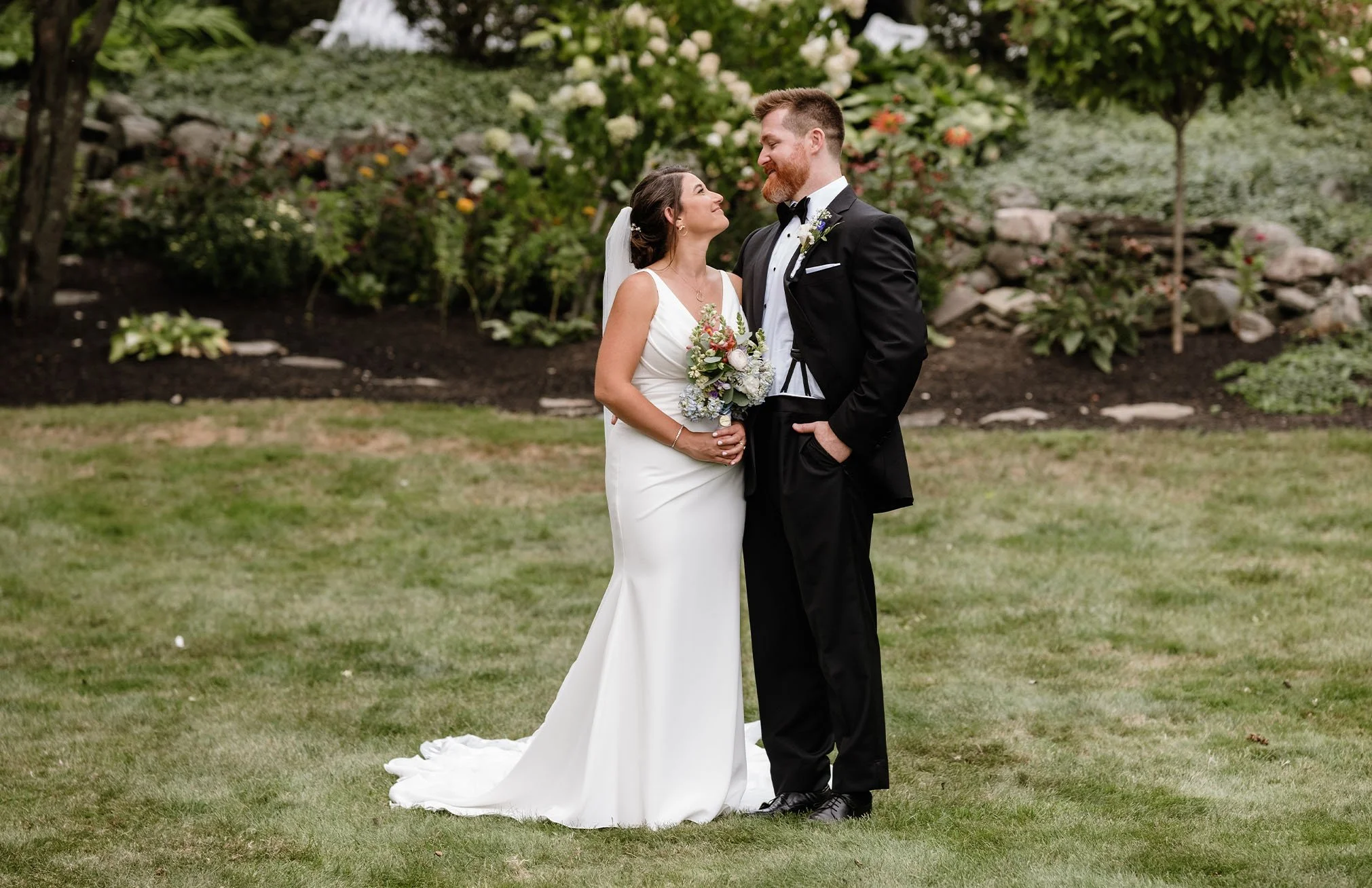 Bride and groom standing outdoors in a garden, smiling and looking at each other, with the bride holding a bouquet of flowers.