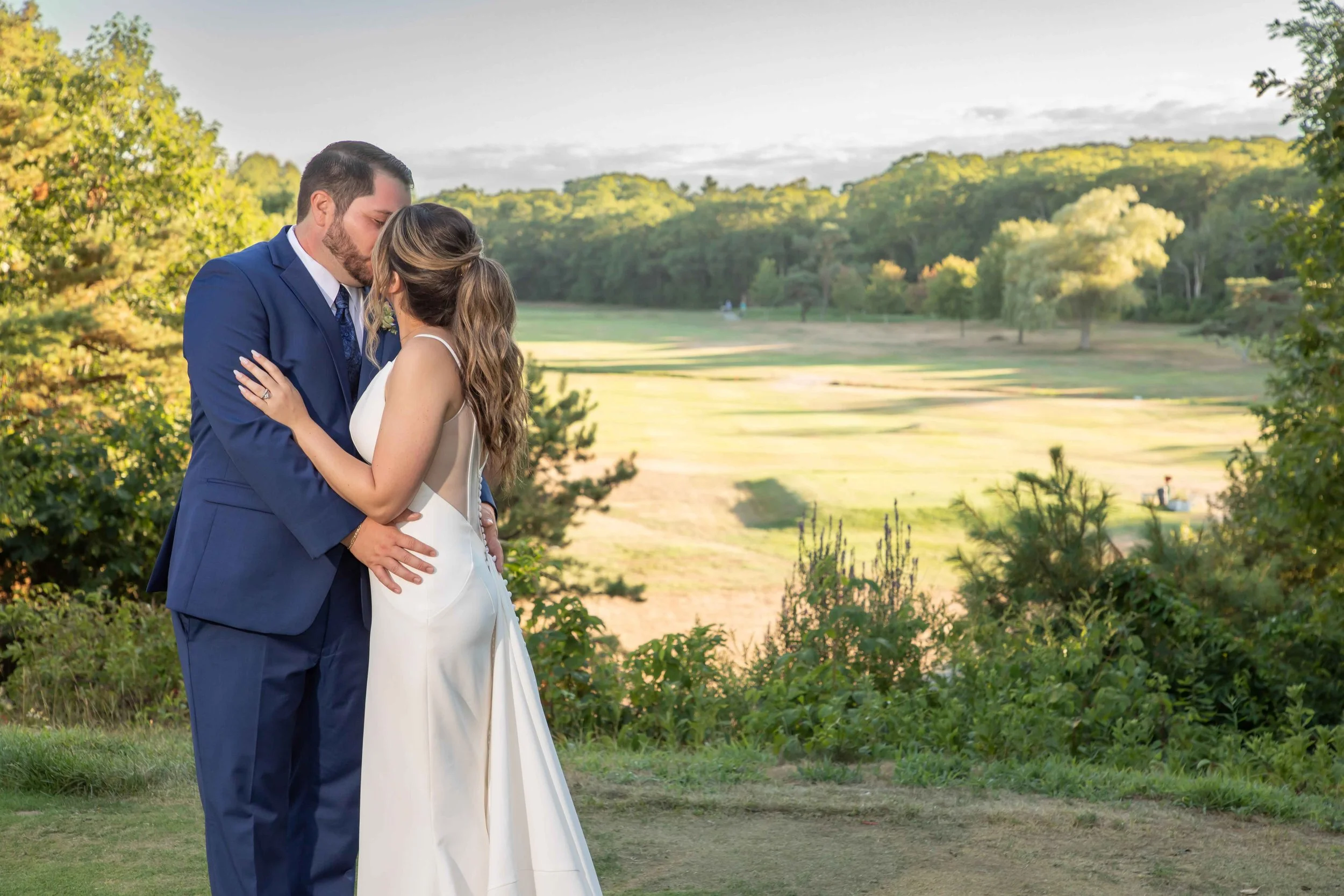 A couple in wedding attire, a man in a blue suit and a woman in a white dress, sharing a kiss outdoors in a green, park-like setting with trees and open fields in the background.