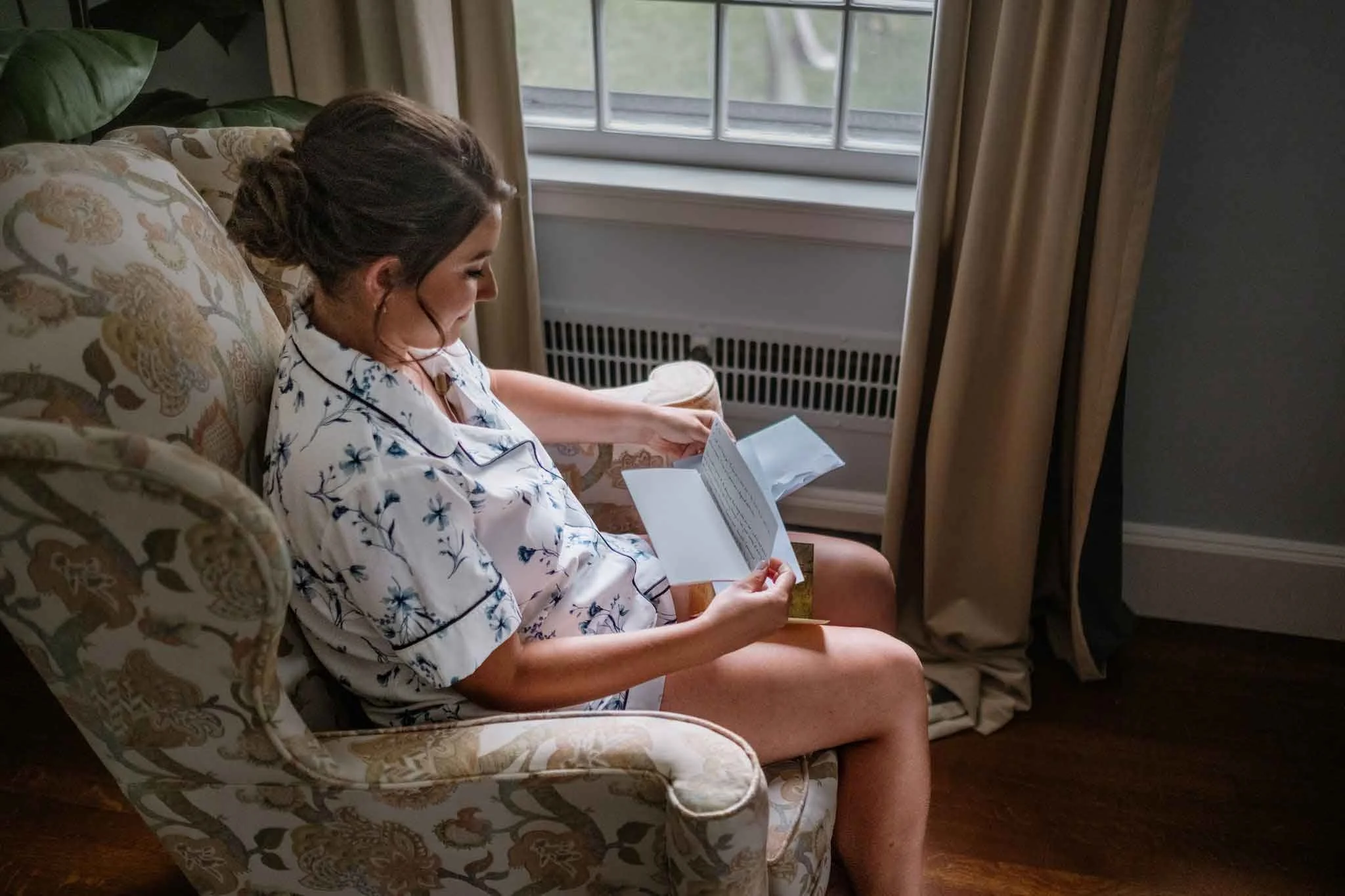 A woman sitting on an upholstered armchair near a window, reading a book, wearing pajamas with a floral pattern.