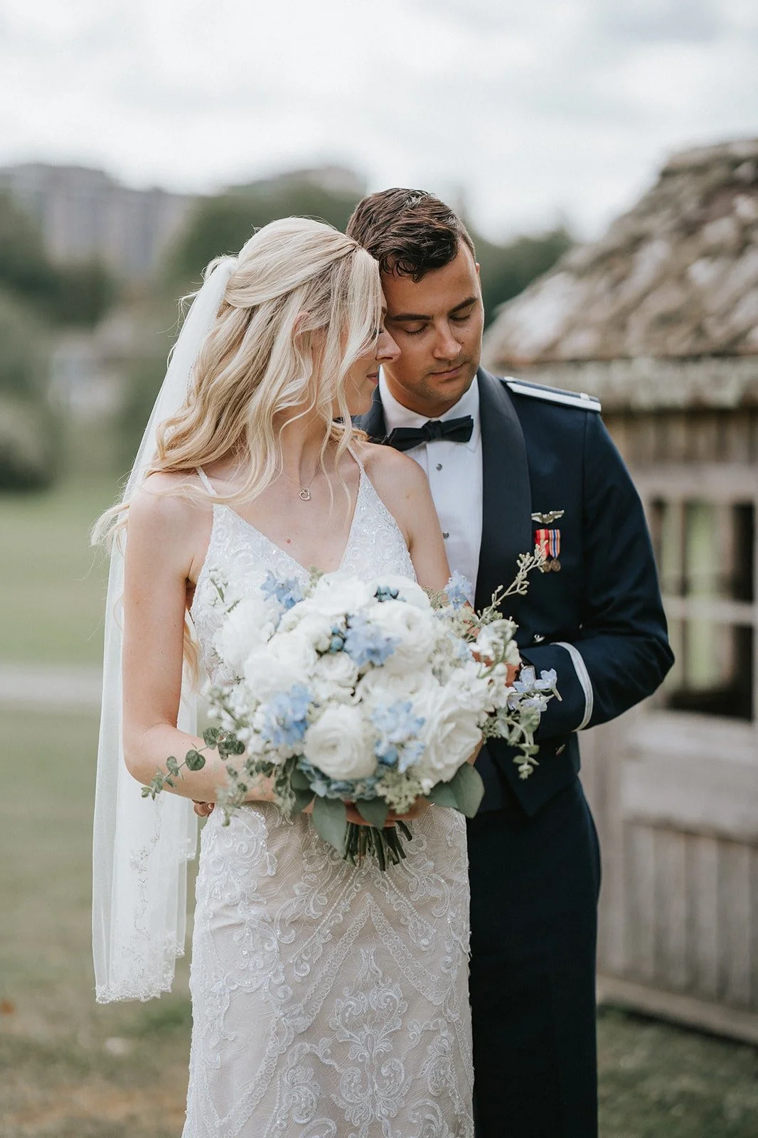 A bride and groom standing close together outdoors, with the bride holding a bouquet of white and light blue flowers, and the groom dressed in a military uniform.