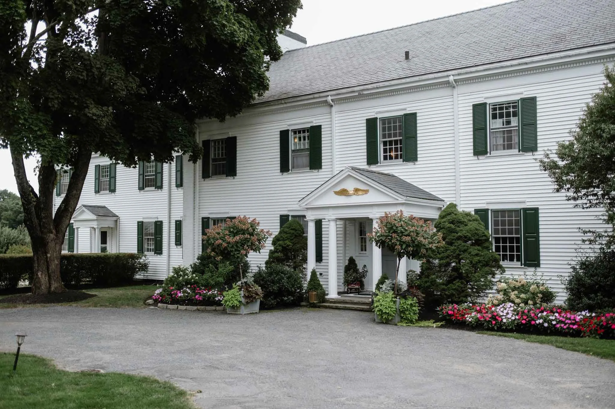 A large white colonial-style house with multiple green-shuttered windows and a centered entryway with a small porch, surrounded by flowering shrubs and trees.