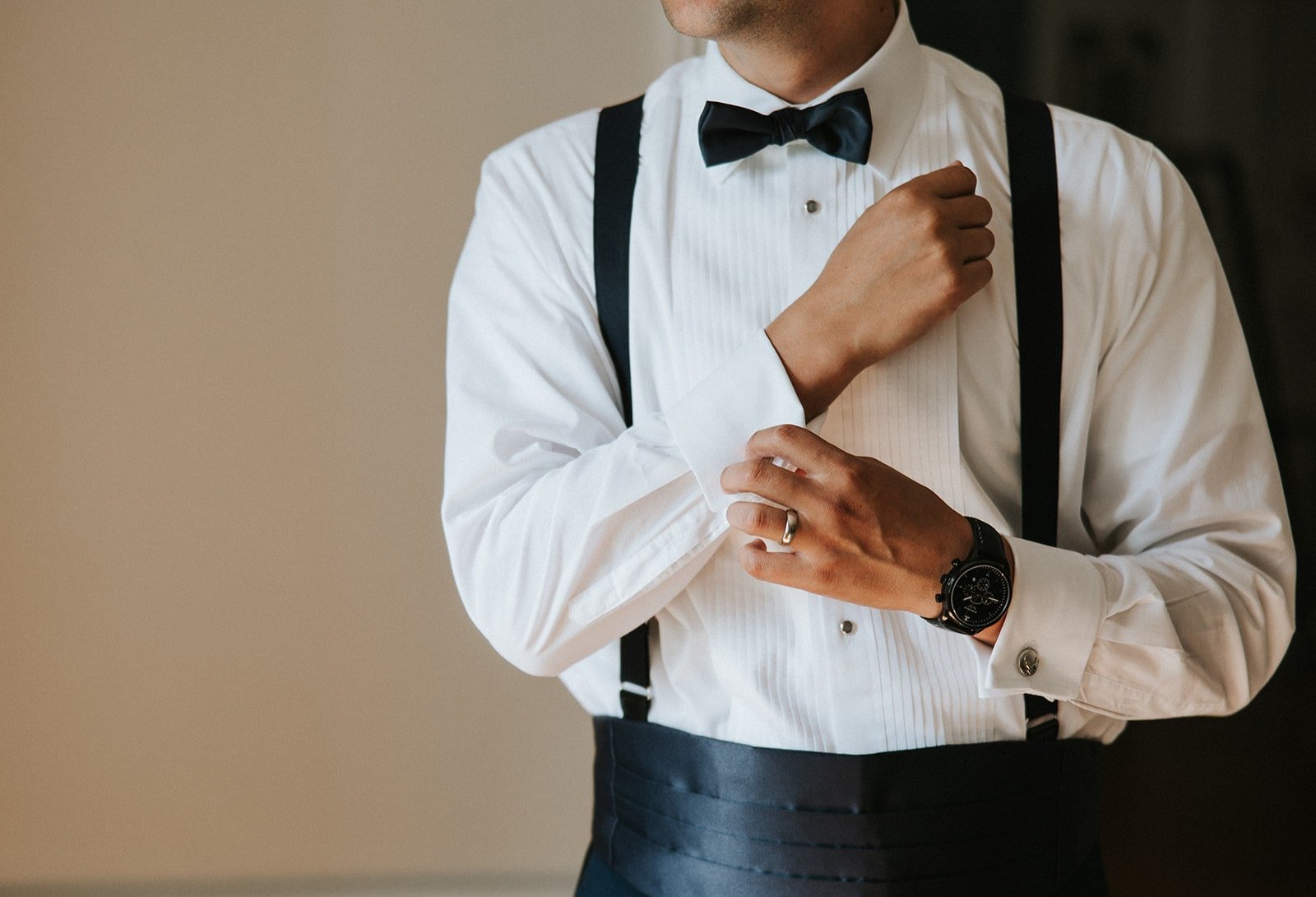 Man adjusting his sleeve, dressed in a white tuxedo shirt with black suspenders, black bow tie, and a watch.