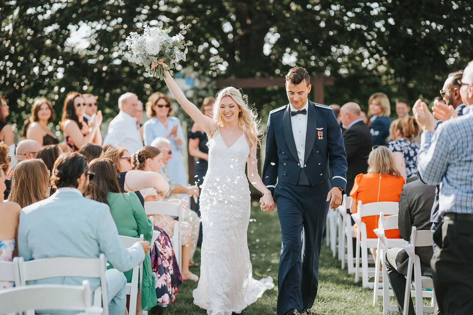 A newlywed couple walking hand-in-hand outdoors during their wedding ceremony, surrounded by seated and standing guests clapping and celebrating, with the bride holding a bouquet and throwing confetti.