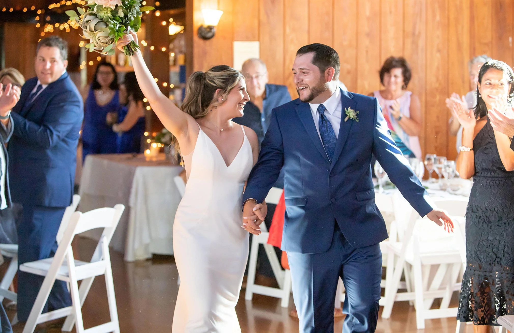 A bride and groom celebrating and holding hands during their wedding reception, surrounded by guests clapping and smiling.