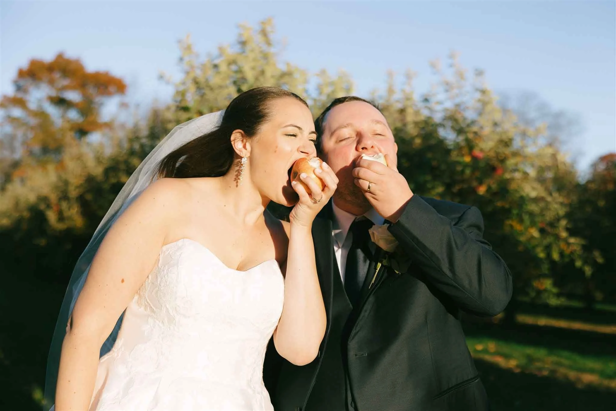 Bride and groom in wedding attire eating cupcakes outdoors in sunlight.