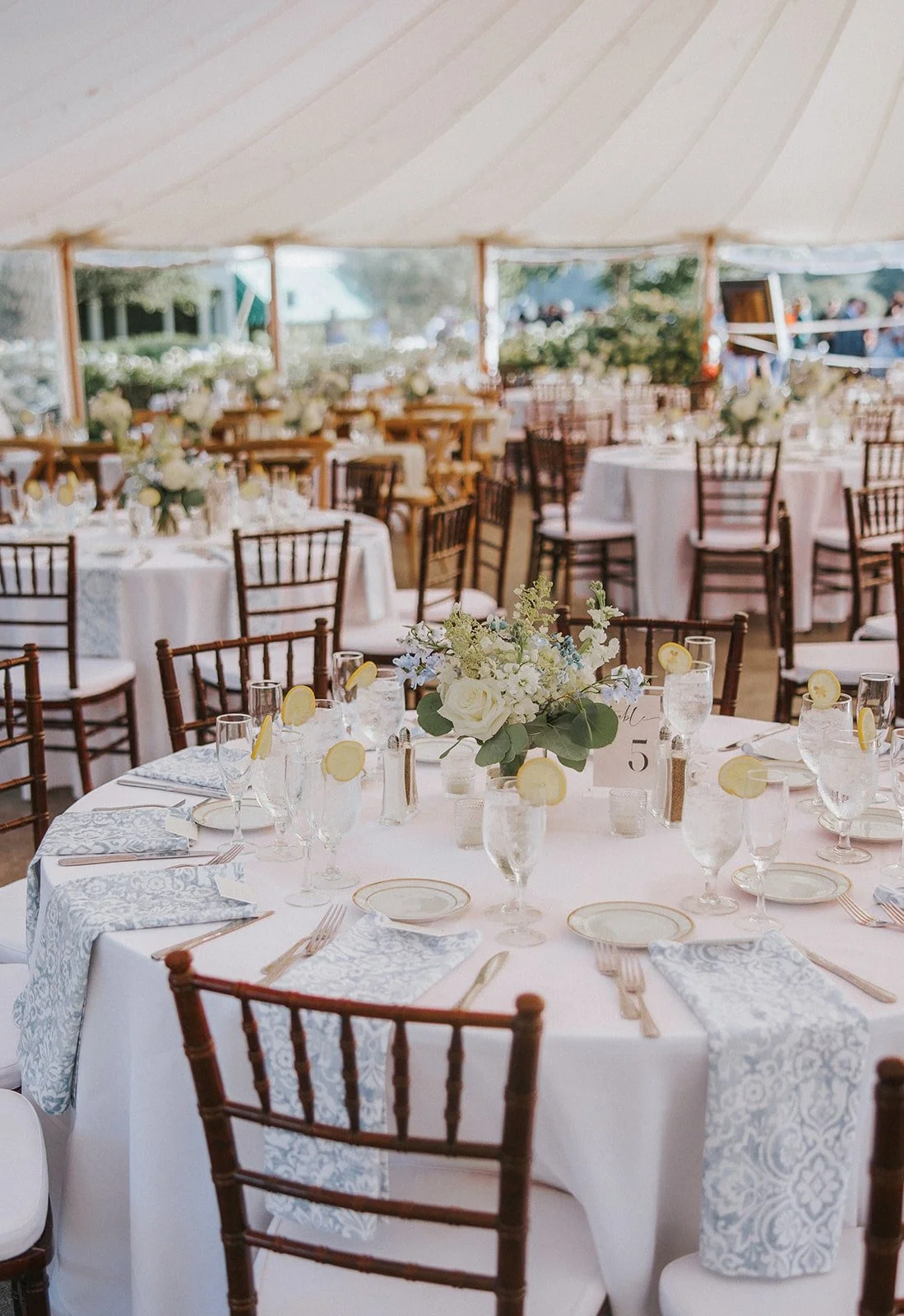 Decorated round wedding reception table under a large white tent with floral centerpiece, napkins, glasses, and silverware, surrounded by wooden chairs.