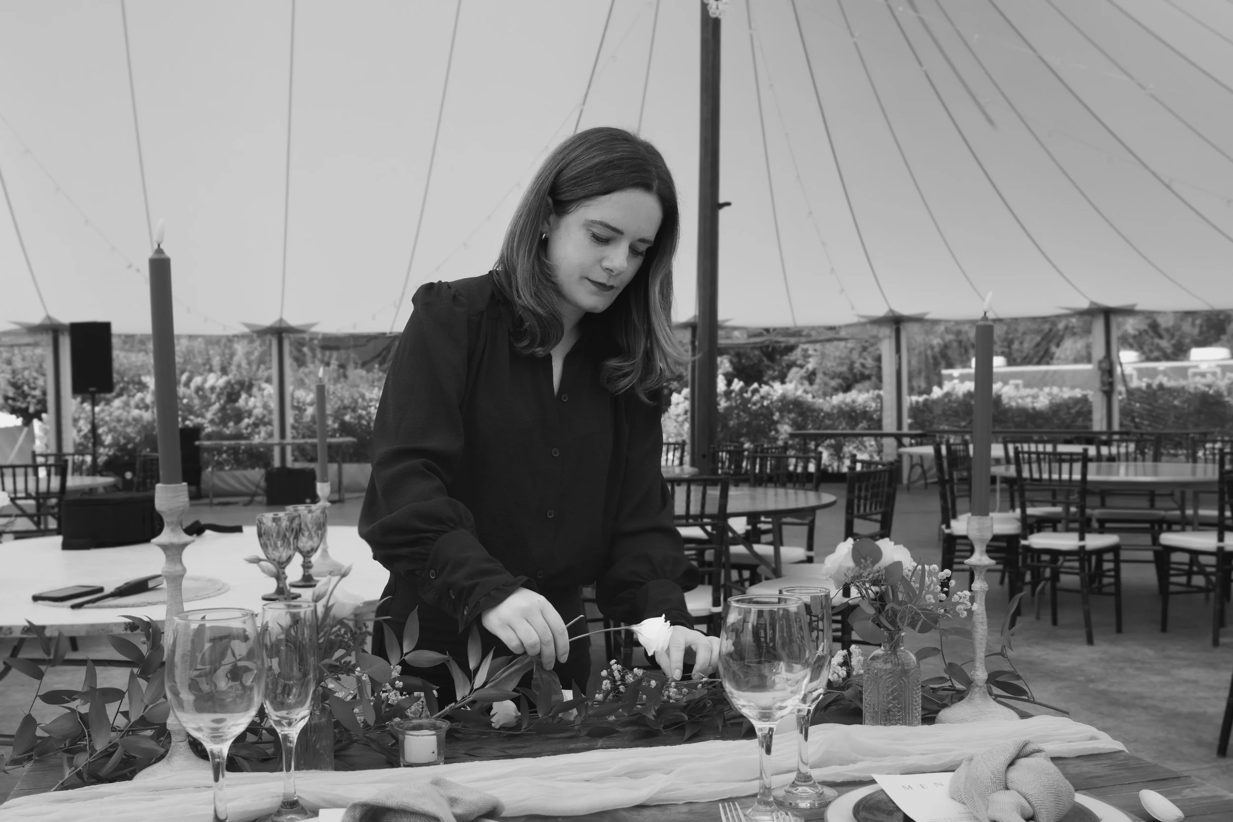 A woman setting up a decorated table under a large tent outdoors, with candles and floral arrangements.