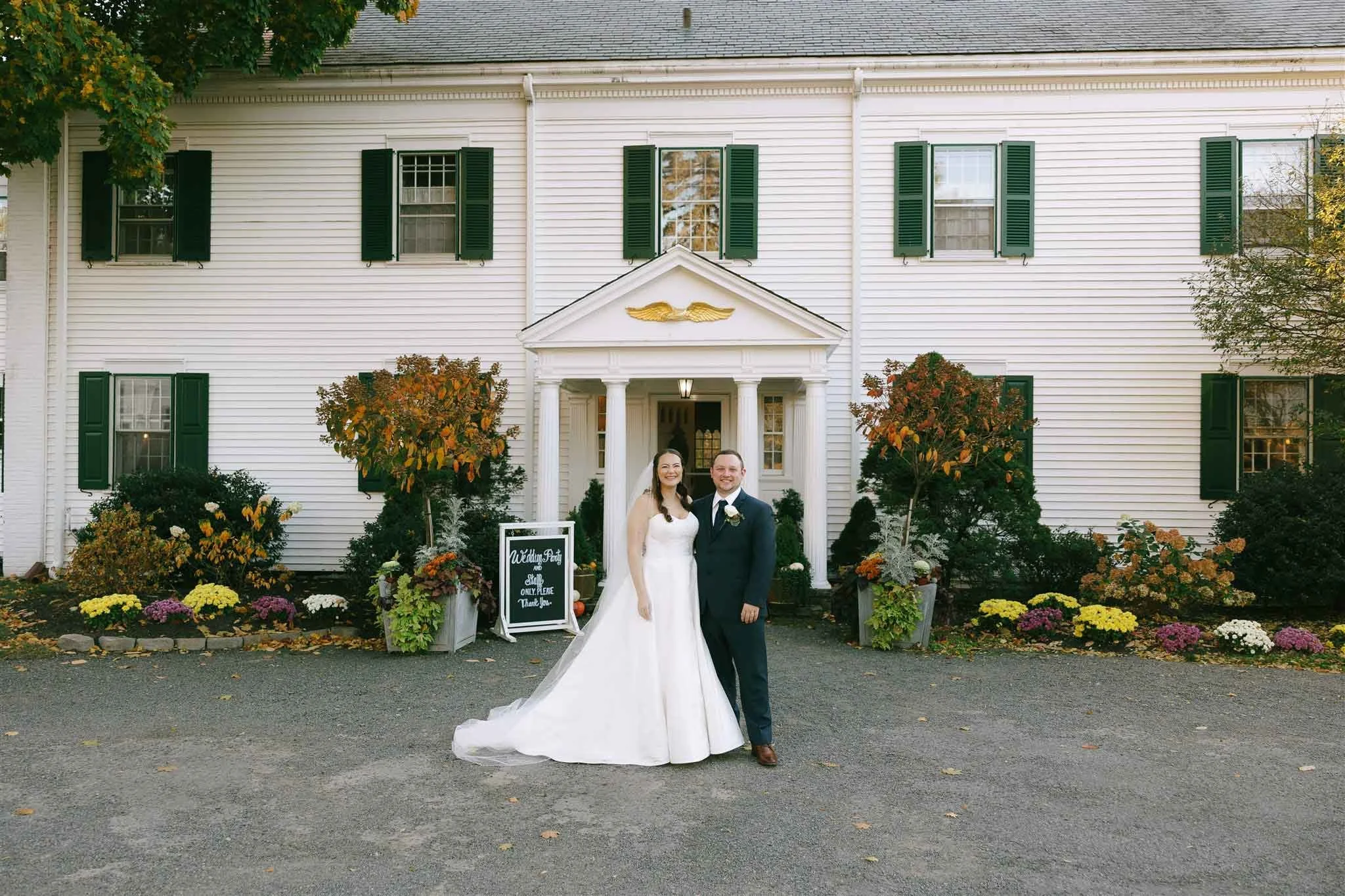 A newlywed couple stands in front of a large white house with green shutters, surrounded by colorful fall flowers and trees. The bride wears a white wedding dress with a long train, and the groom wears a dark suit. There is a wedding sign near them.