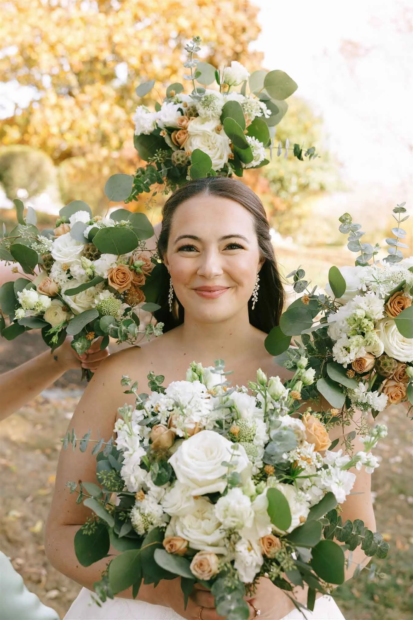 A smiling woman holding a bouquet of white and peach flowers, surrounded by large floral arrangements, outdoors during fall with trees in the background.