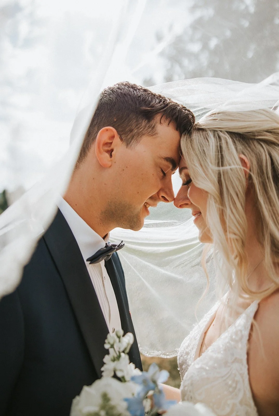 A newlywed couple standing close with their foreheads touching, smiling with eyes closed under a sheer white veil.