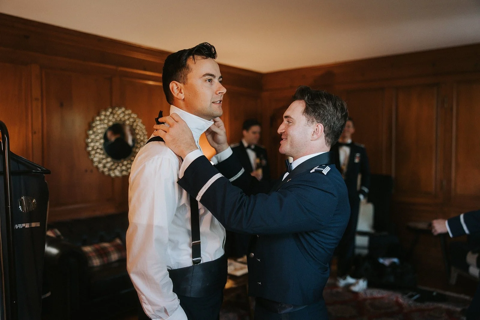A man in a white shirt and suspenders is being assisted by another man in a dark military uniform as he adjusts his collar, with other men in uniform in the background inside a wood-paneled room.