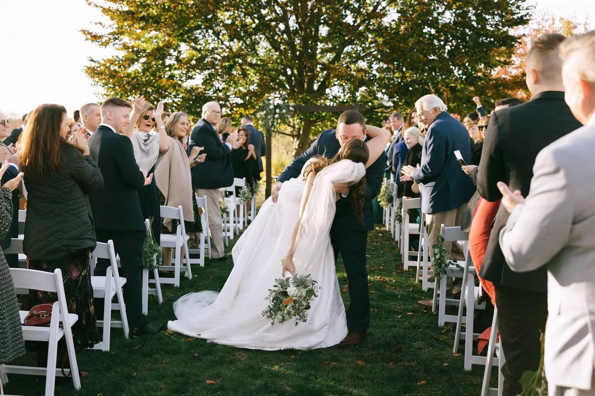 Bride and groom kissing during an outdoor wedding ceremony with guests clapping and cheering.
