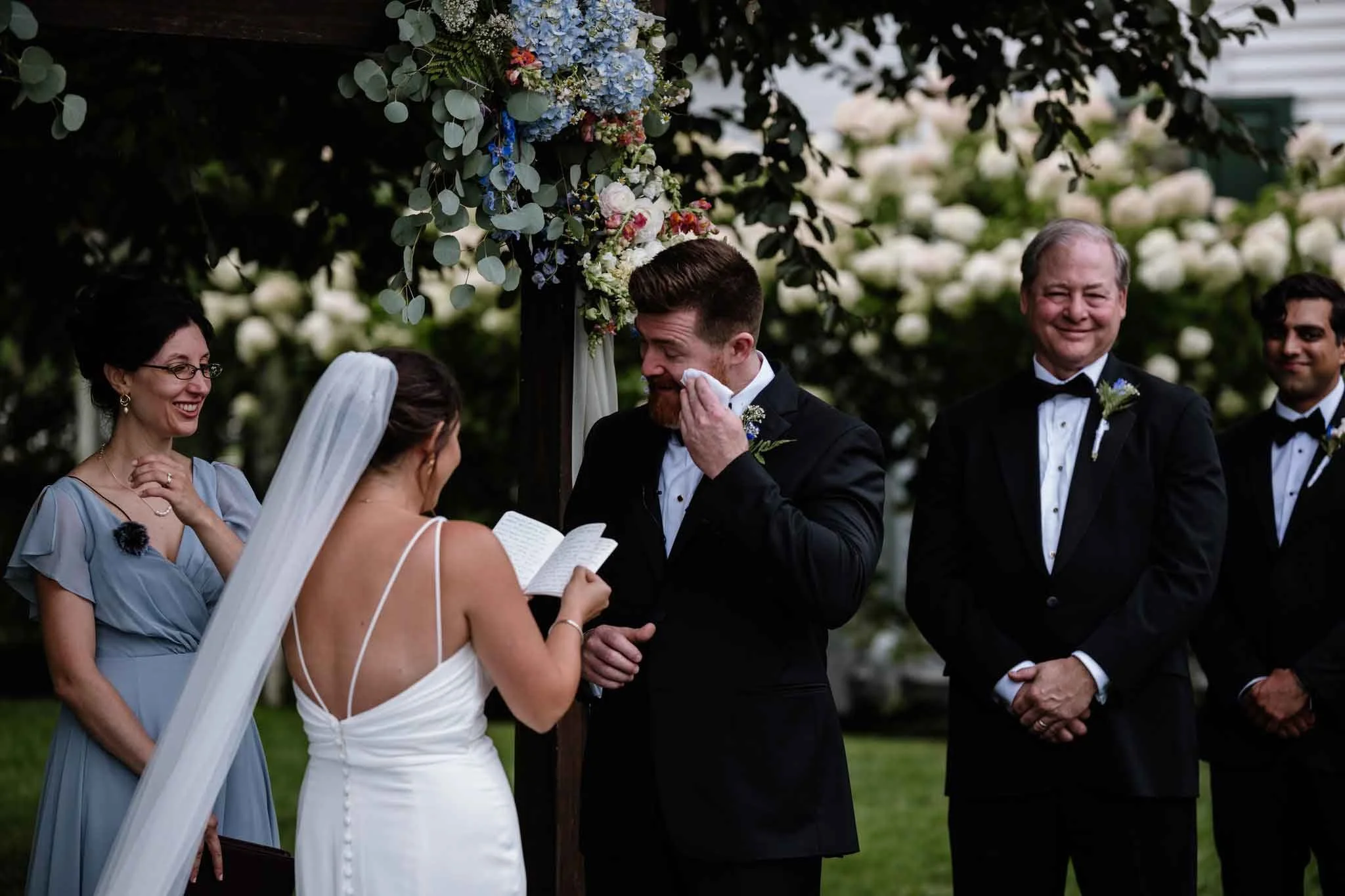 A wedding ceremony with the bride and groom exchanging vows outdoors under a floral arch, surrounded by friends and family, including a woman in a light blue dress and men in black tuxedos.
